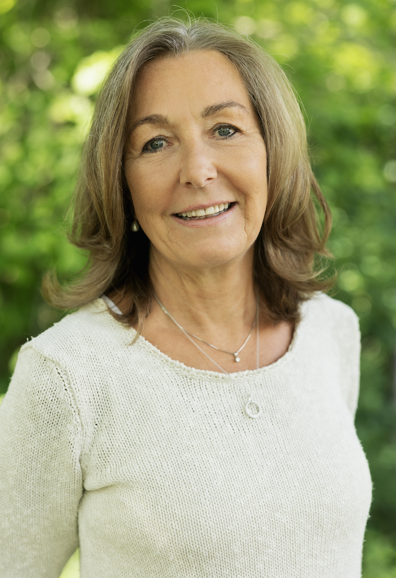 Portrett av Cathrine Berge, grunnleggeren av God Helse AS. Hun har på seg en hvit genser, smiler inn i kameraet og i bakgrunnen er det grønne trær i bokeh.