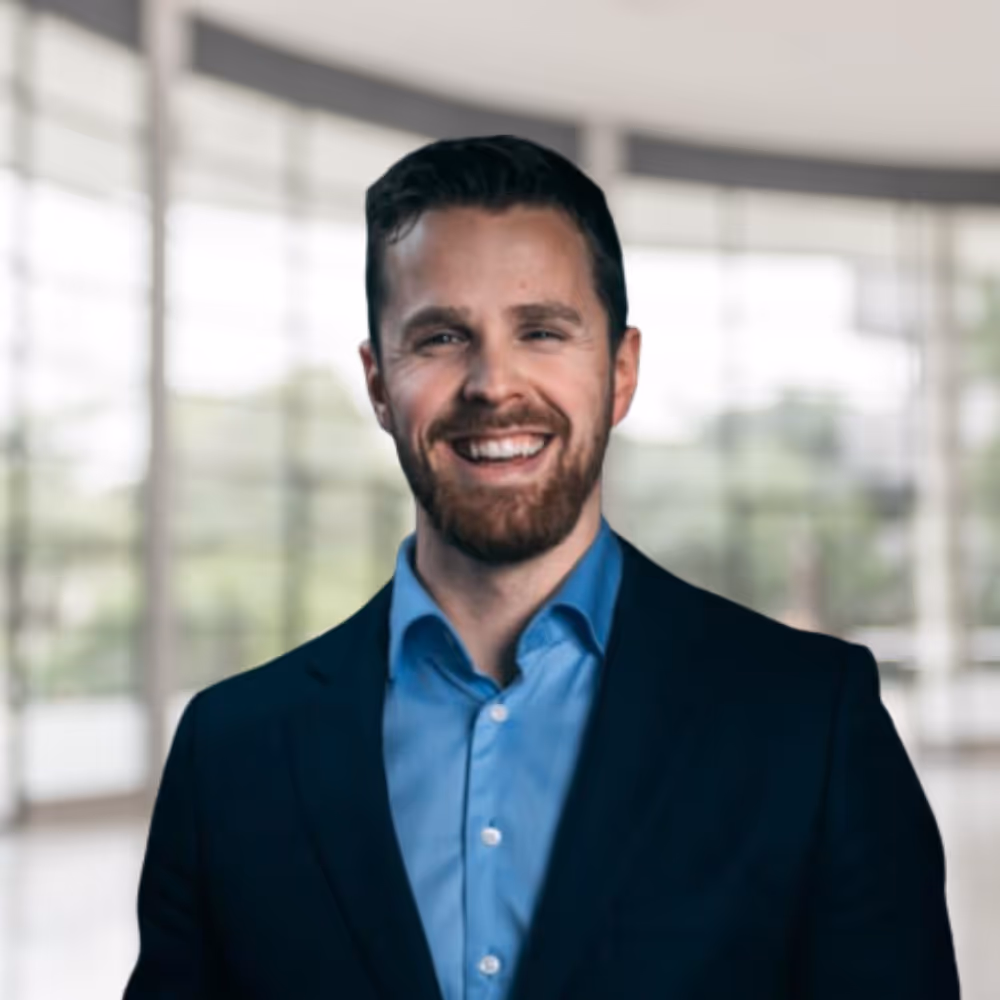 Smilende mann med kort brunt hår og skjegg iført blå skjorte og mørk blazer foran uskarpt kontormiljø.