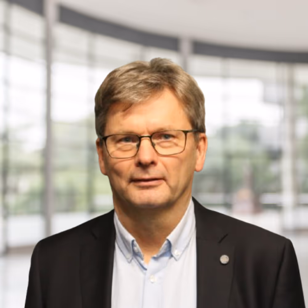 Mann med briller og grått hår iført hvit skjorte og svart jakke foran uskarpt kontormiljø.