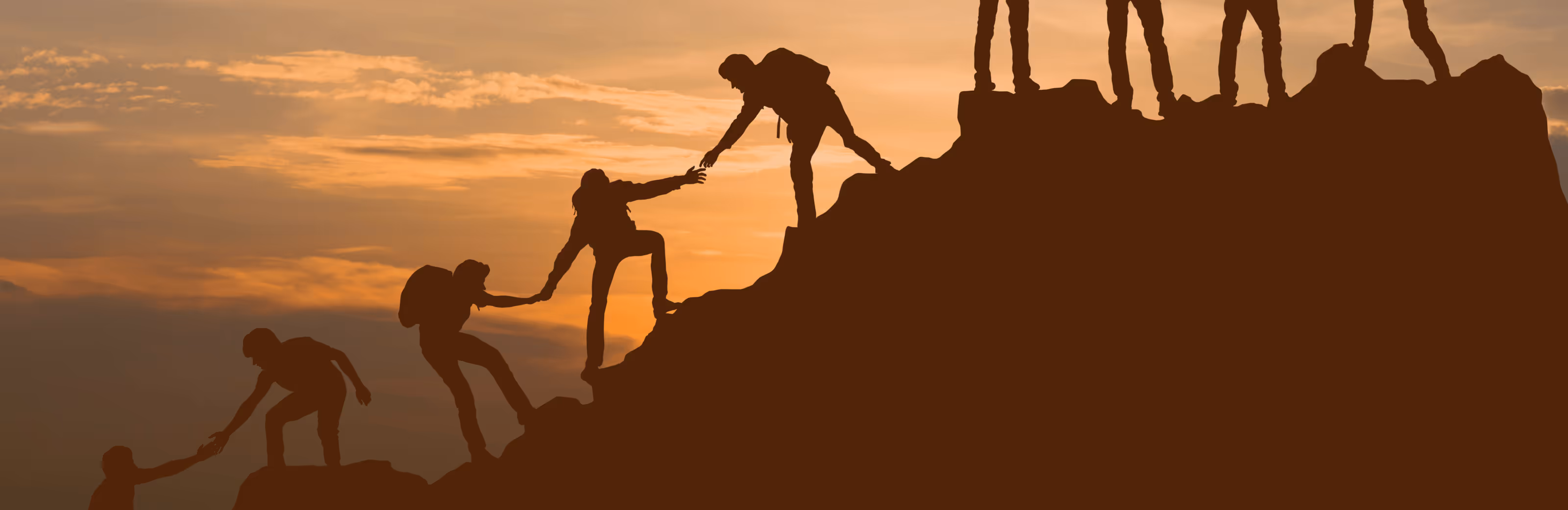 Silhuetter av folk som hjelper hverandre opp en fjellside under solnedgangen.