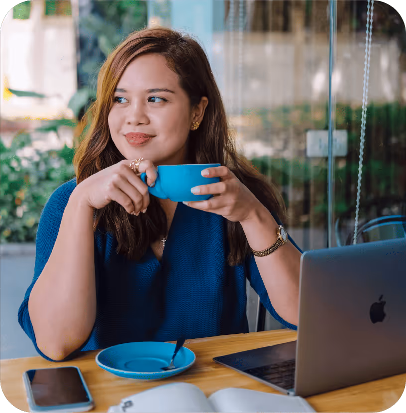 Woman sitting at a café table, holding a blue coffee cup while working on a laptop, with a phone and notebook nearby.