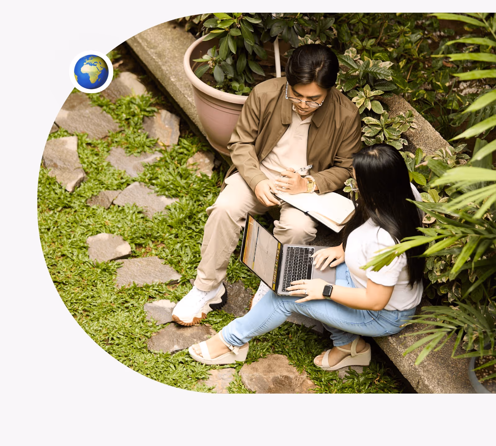 Two students sitting outdoors on a garden pathway, collaborating with a laptop and notebook surrounded by greenery and plants.
