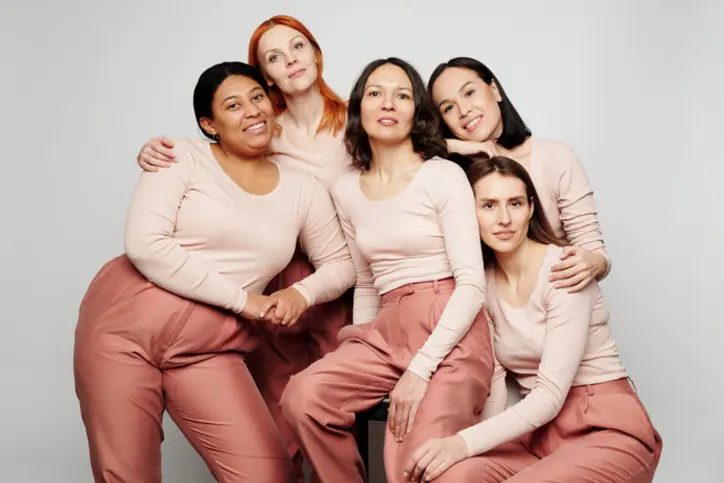 Five women of diverse backgrounds wearing matching pink tops and pants posing closely together against a plain light gray background.