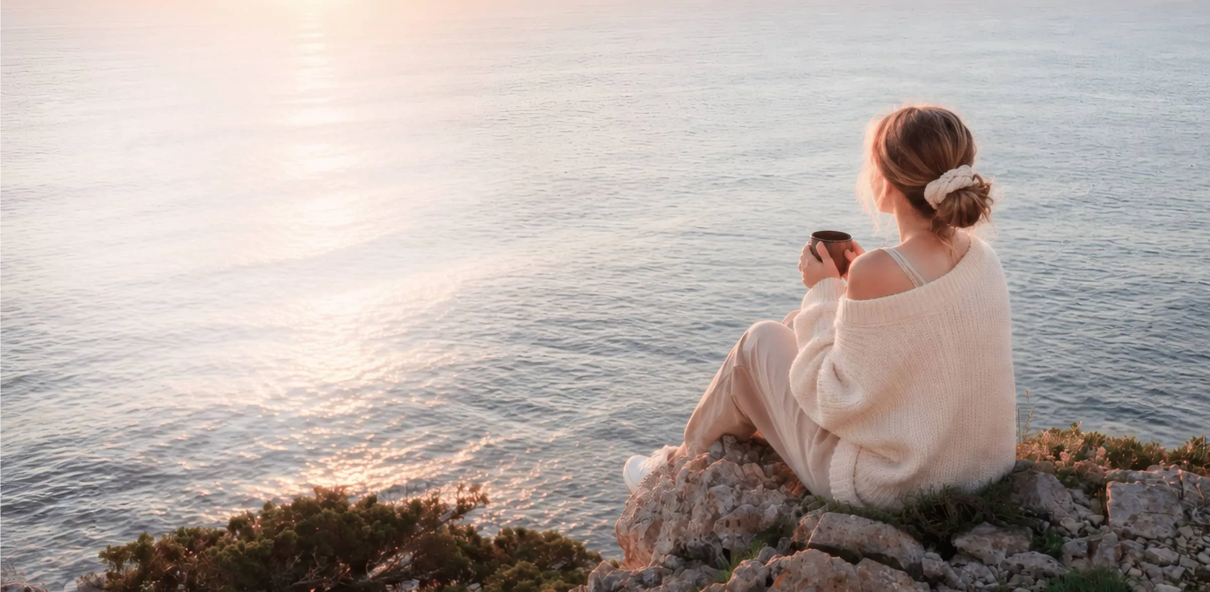 A woman wearing a white off-shoulder sweater and beige pants sits on rocky terrain near water, holding a cup and gazing at the calm ocean during sunset.