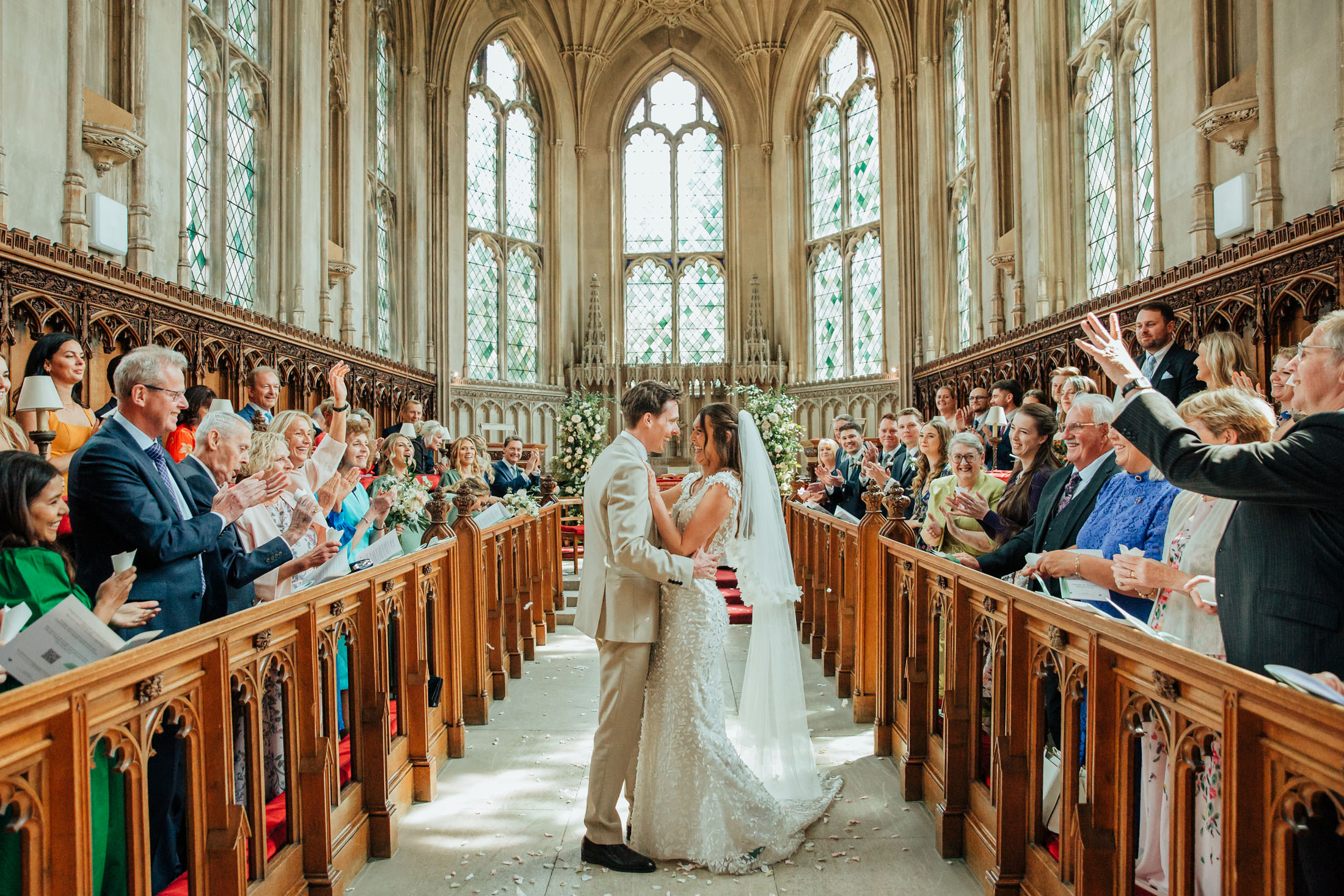 Bride and groom embracing in a church aisle with guests clapping and cheering on both sides at Ashridge House Chapel in Hertfordshire.