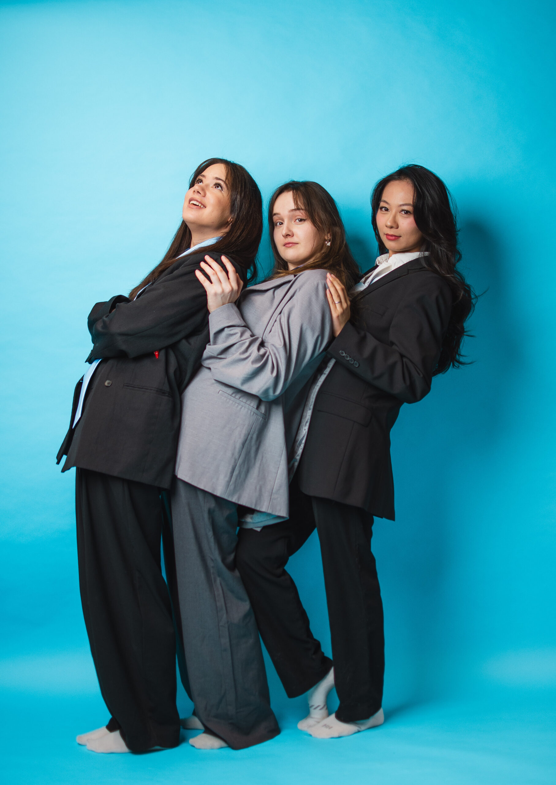 Three women, viewed from the left, in front of one another, standing in front of a turquoise background.