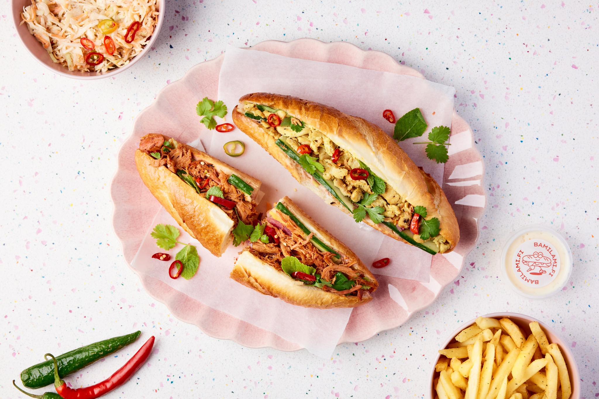 Vietnamese sandwiches on a pink plate. Fries in a bowl on the bottom right. Salad in a bowl on the top left. Two jalapeño peppers on the bottom left.