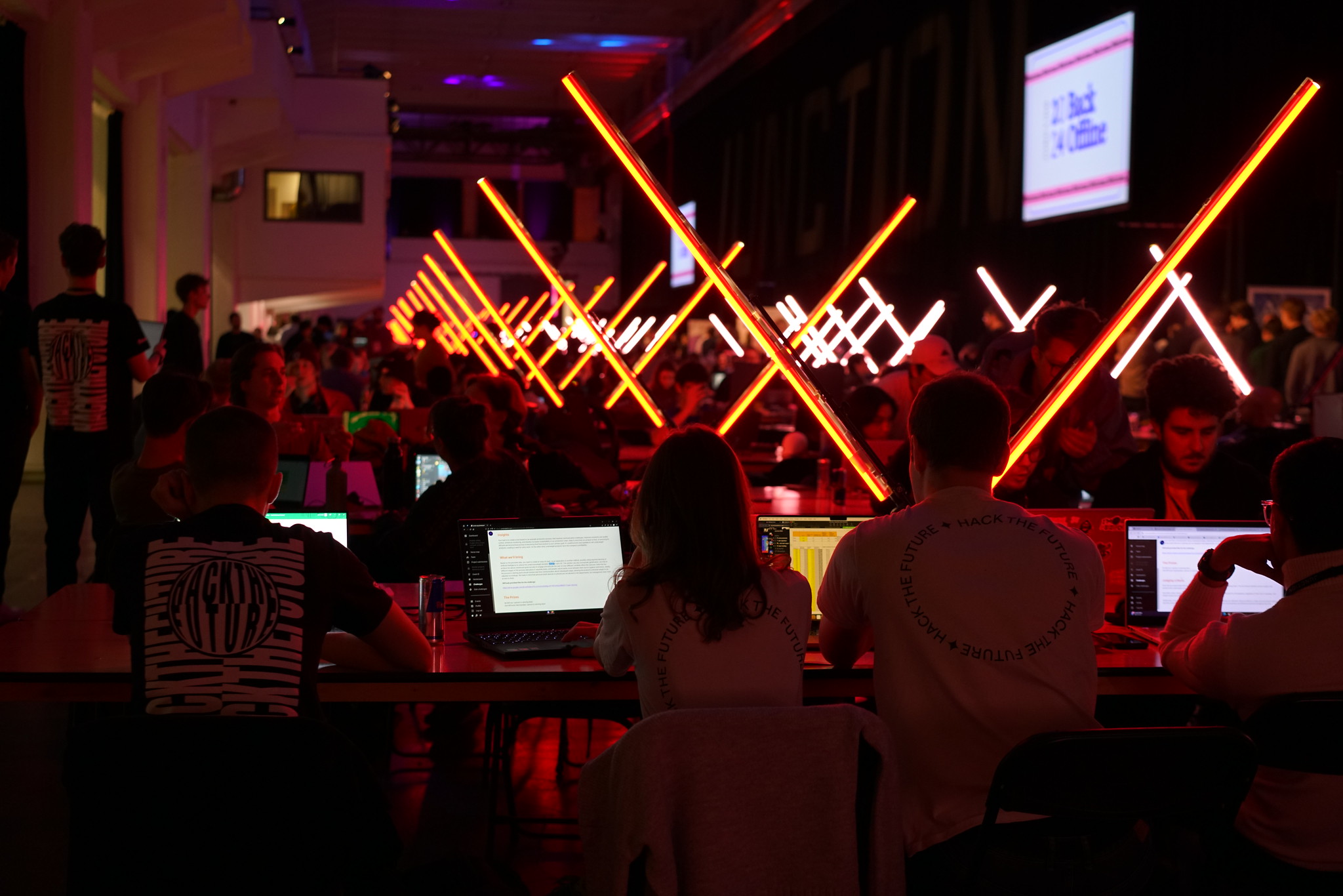 Picture from a Junction hackathon event. Lighting is dim, people are sitting at tables at their laptops, neon signs separate the tables.