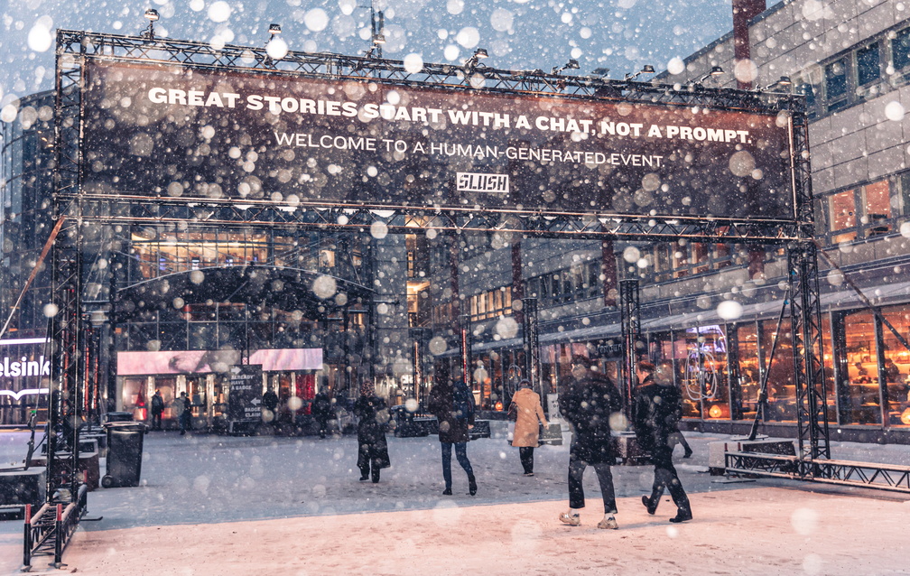 Slush 2024 event entrance. Banner says: Great stories start with a chat, not a prompt. Welcome to a human-generated event. People are walking into the venue. Snow is falling.