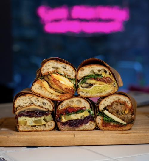 Five sandwiches on a wood table in front of a dark blue wall with a pink neon sign