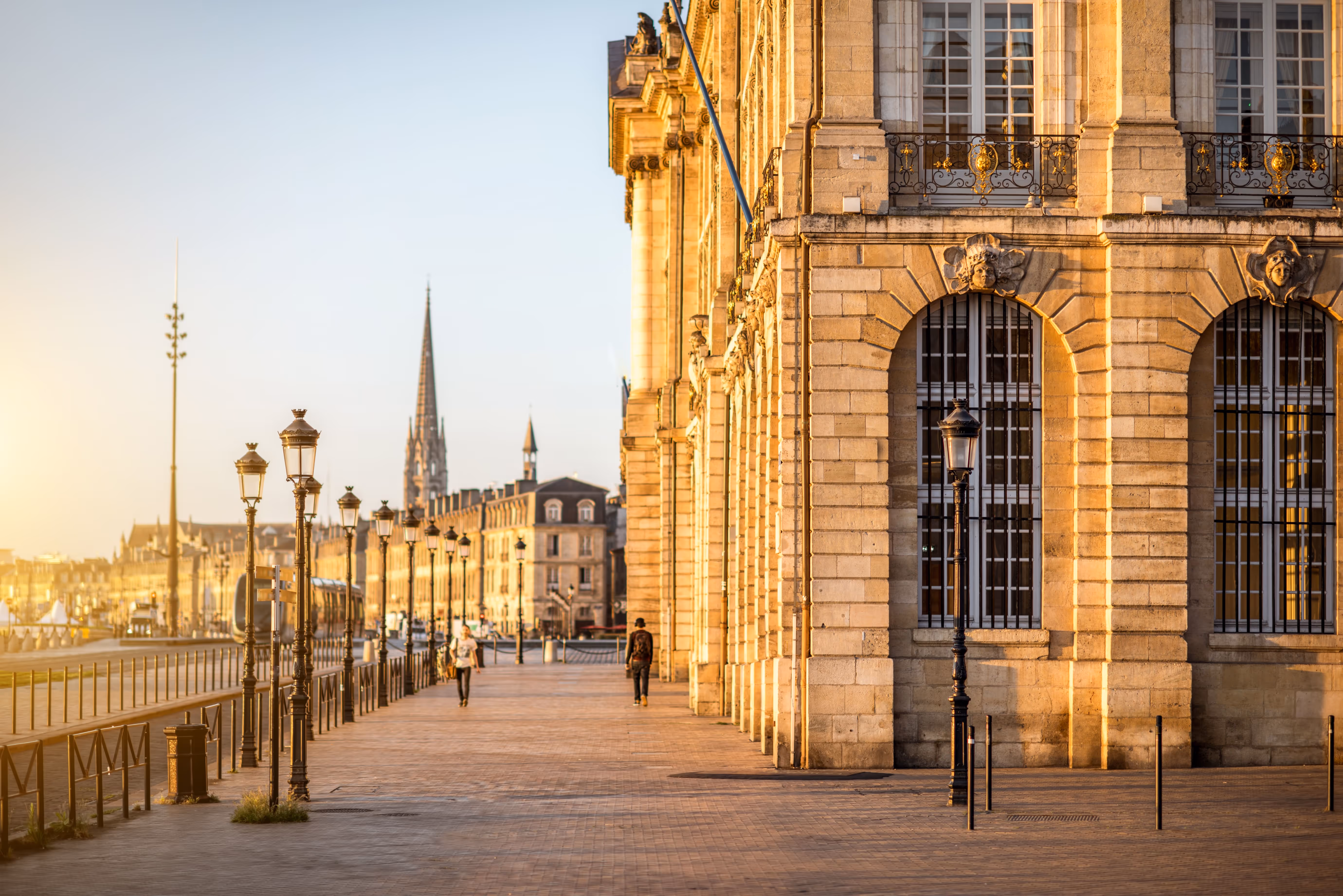 Vue ensoleillée des quais de Bordeaux avec des lampadaires anciens, des passants et la façade d’un bâtiment historique en pierre, sur fond de ciel dégagé et de la flèche de l’église Saint-Michel à l’horizon.