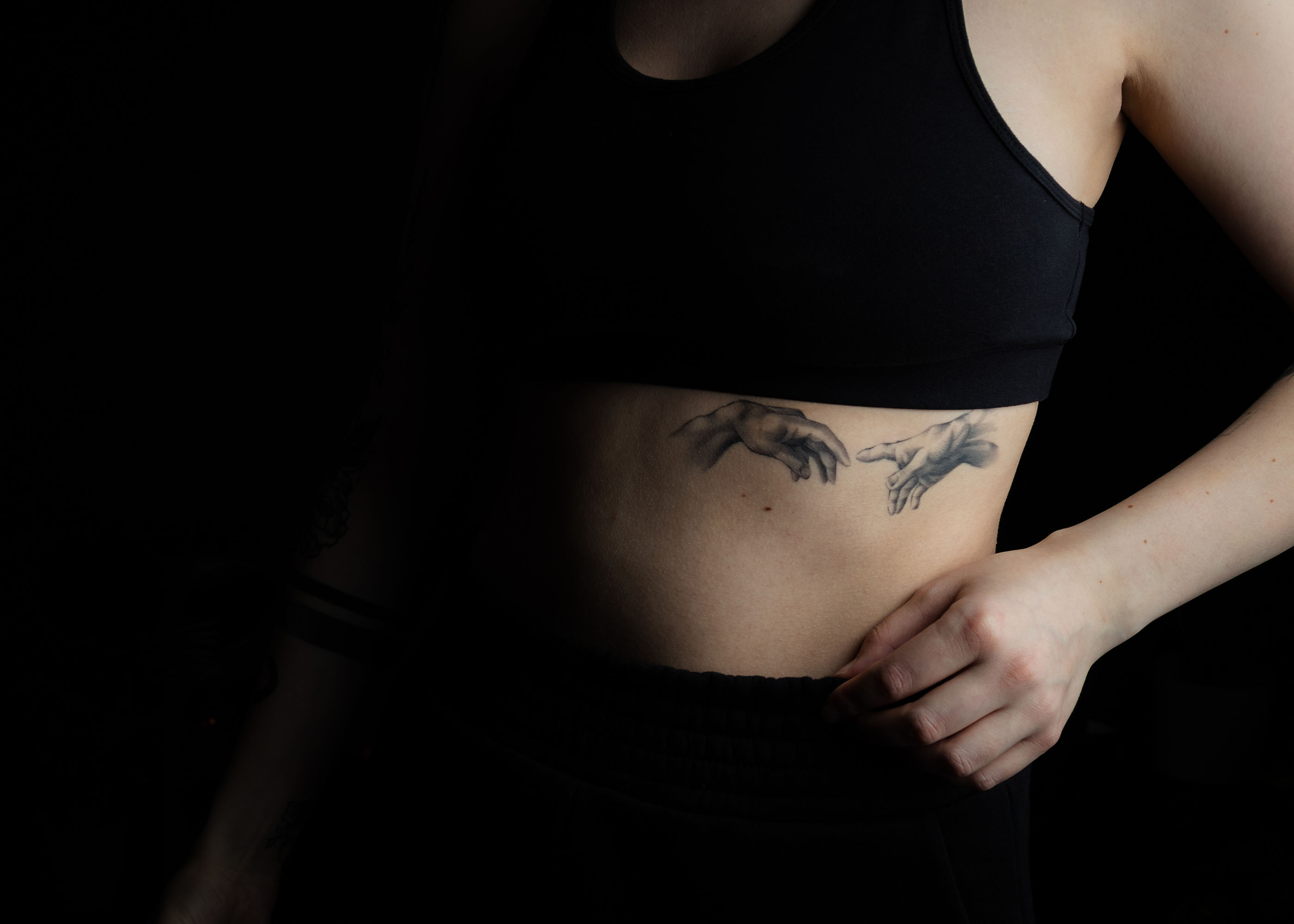 Tattoo mit zwei sich fast berührenden Händen auf dem seitlichen Bauch einer Person mit schwarzem Oberteil.