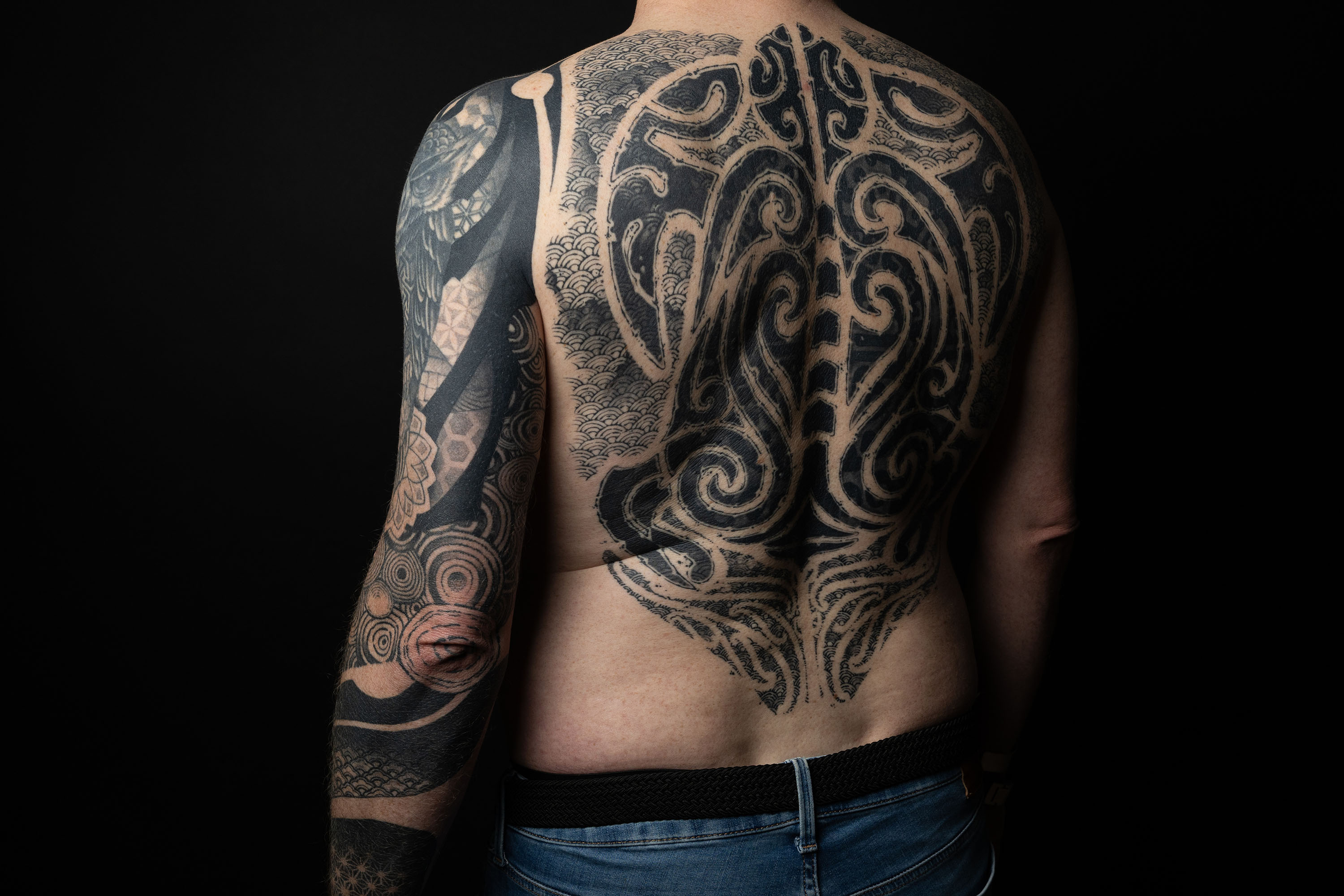Rückenansicht eines Mannes mit auffälligem, großflächigem schwarzen Maori-Tattoo auf Rücken und linkem Arm vor schwarzem Hintergrund.