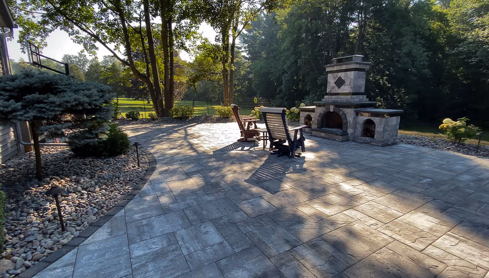 A curved paver patio in Whitesboro, NY with a grand stone fireplace and two adirondack chairs