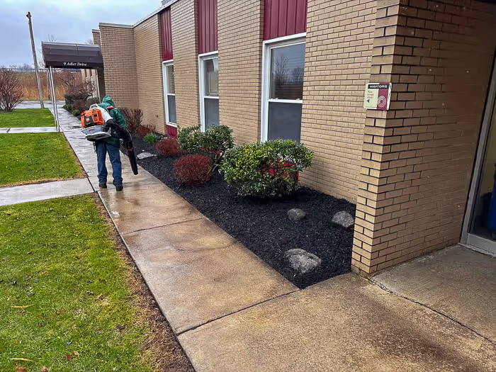 A Pro Snow Removal employee using a leaf blower on a wet sidewalk and flower bed