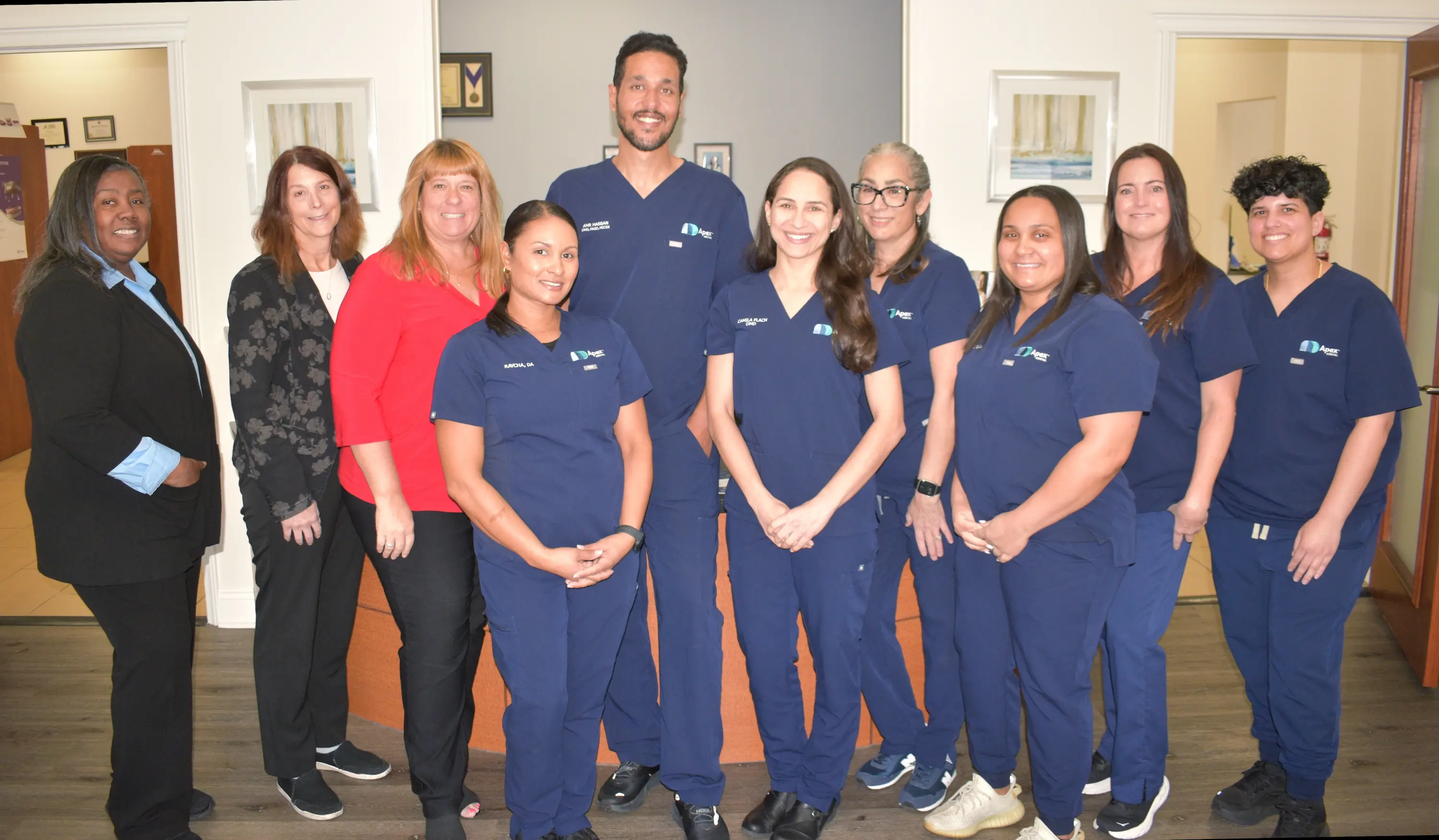 Group photo of ten healthcare professionals and office staff standing indoors, most wearing navy blue medical scrubs and smiling.