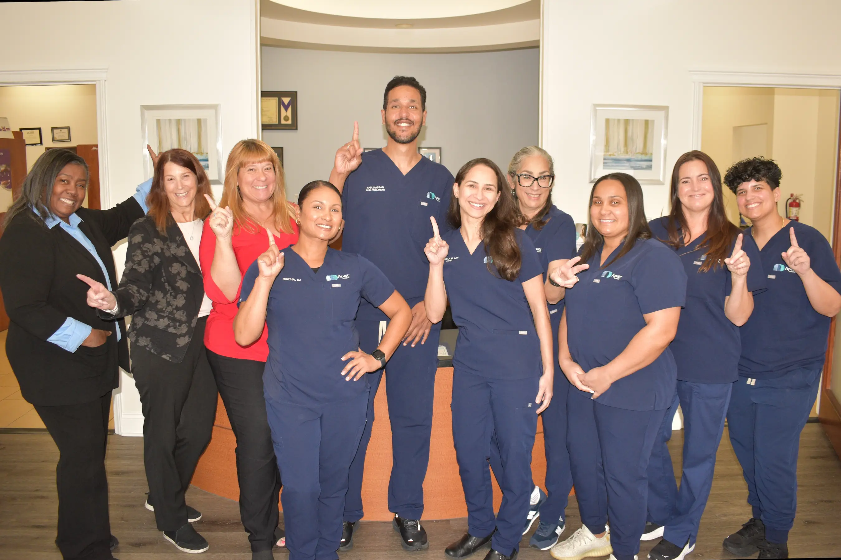Group of ten smiling medical staff and office workers posing indoors, many wearing navy blue scrubs and holding up one finger.