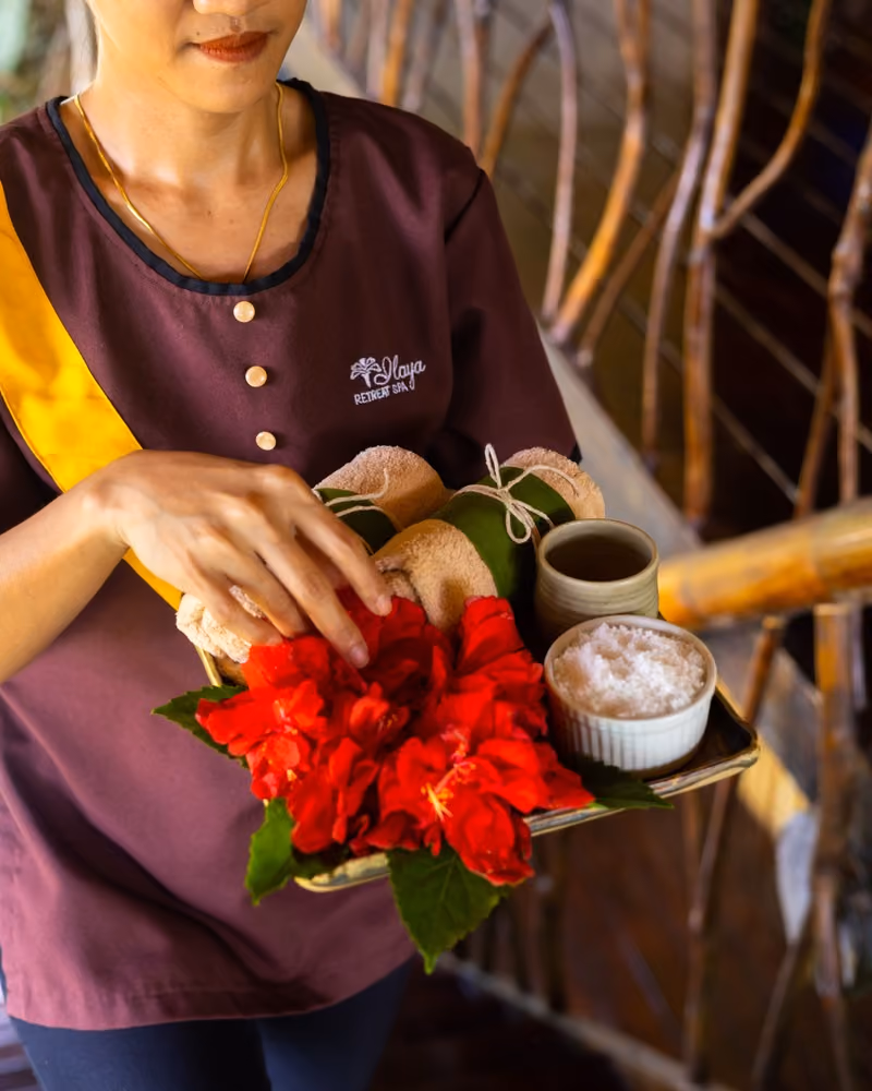 Spa therapist at Ilaya Retreat Spa holding a tray with red hibiscus flowers, towels, and herbal wellness items for a traditional Filipino spa treatment.