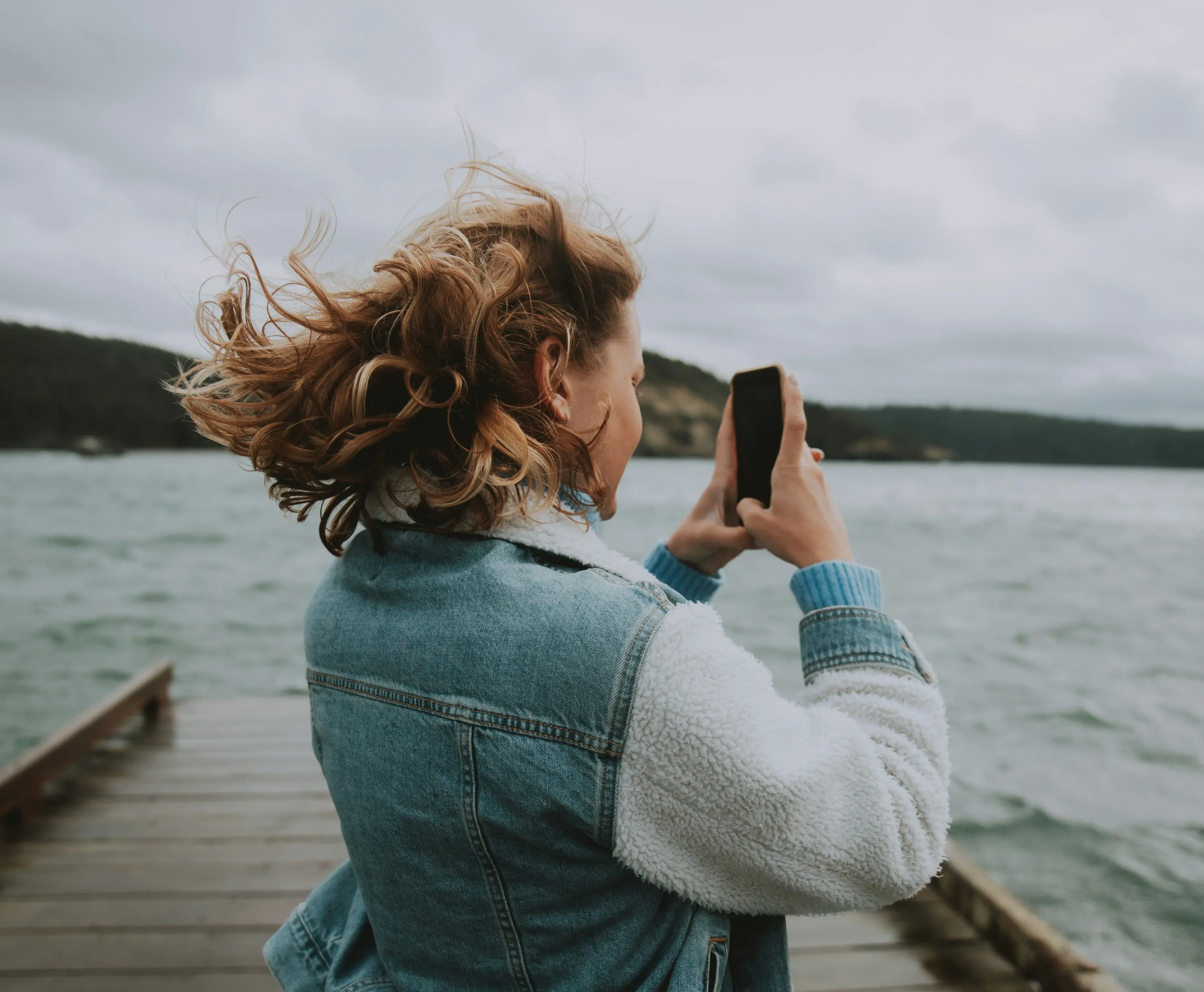 Woman standing on a dock taking a photo of the lake with her phone on a windy day.
