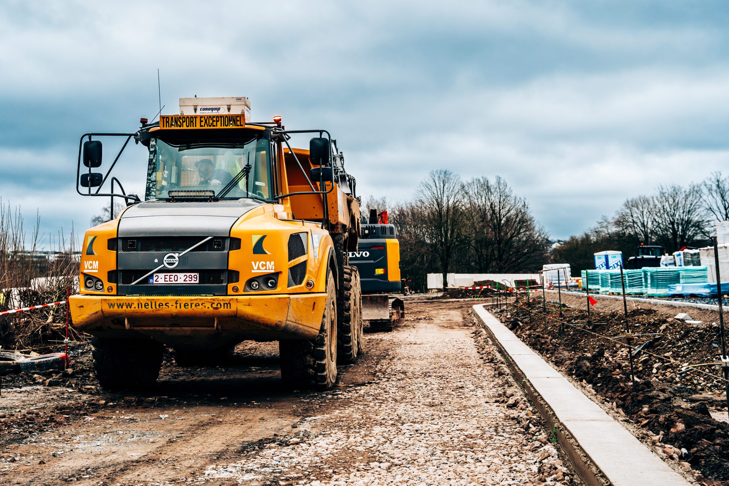 Chantier en cours pour le projet immobilier 'Clos des Arts' : engin de construction Volvo préparant le terrain pour les futures maisons neuves.