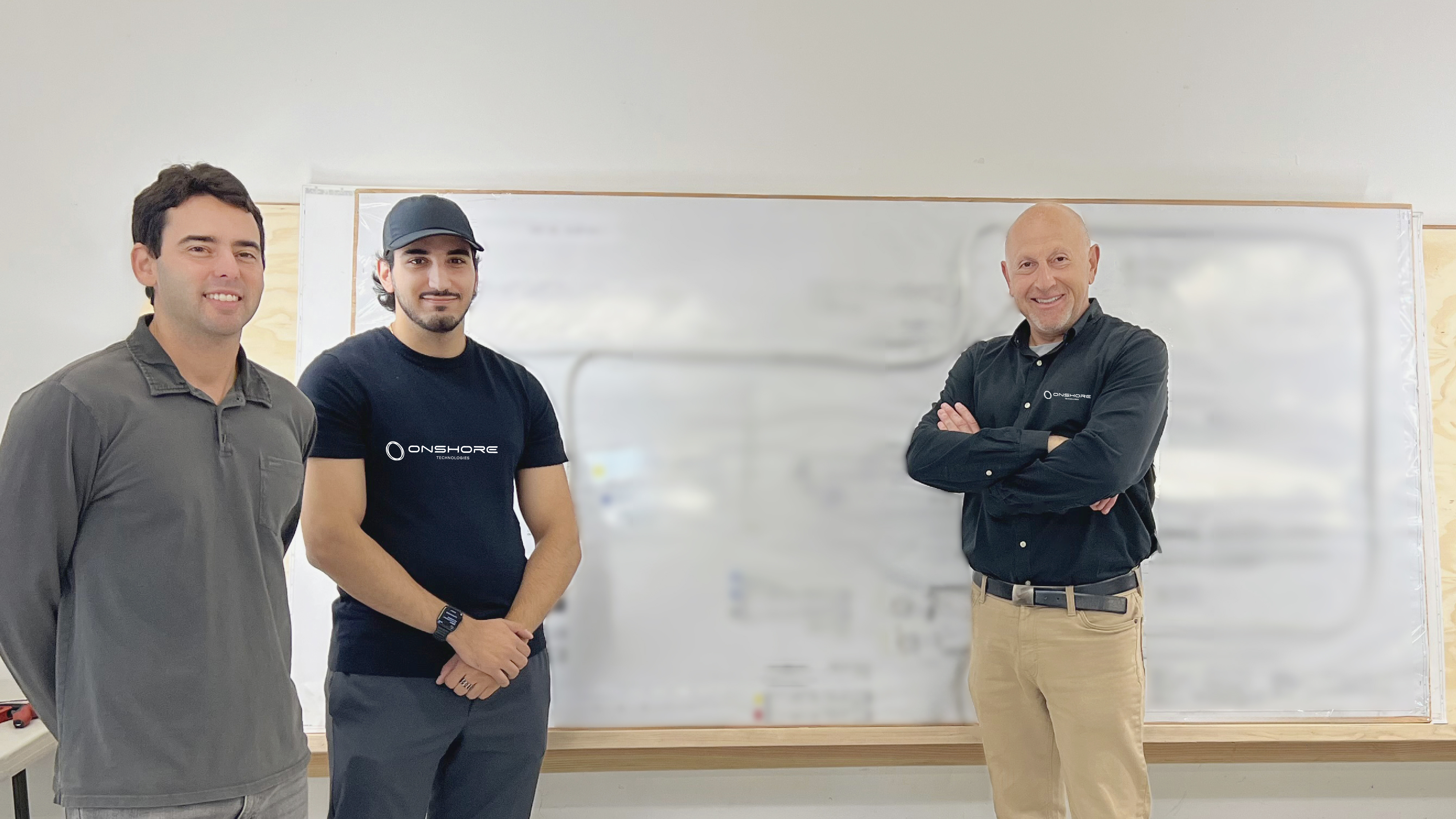 Three men standing and smiling in front of a large engineering blueprint board with wiring diagrams.