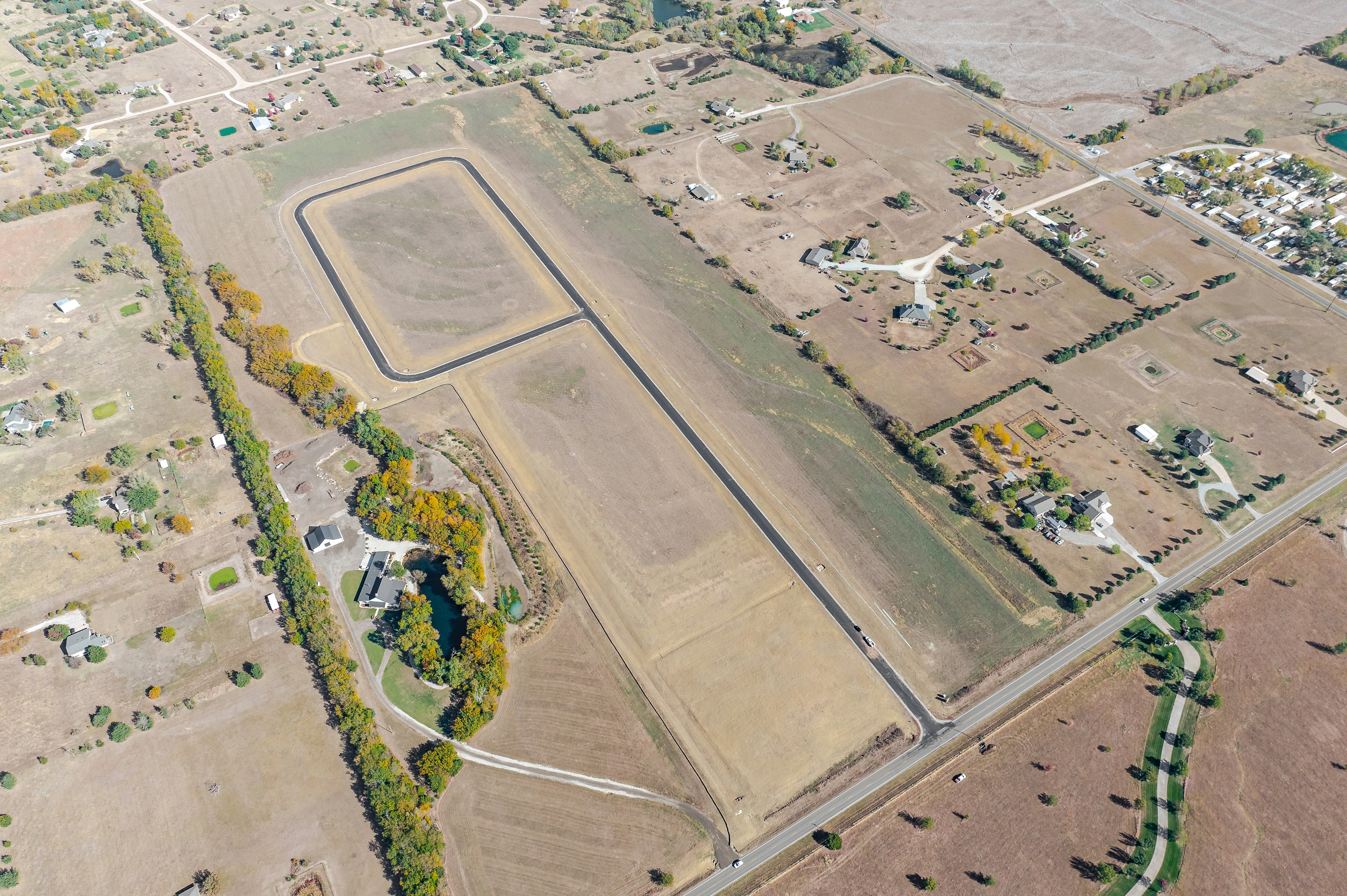 Aerial view of a rural landscape with a rectangular paved track, scattered houses, fields, trees, and dirt roads.