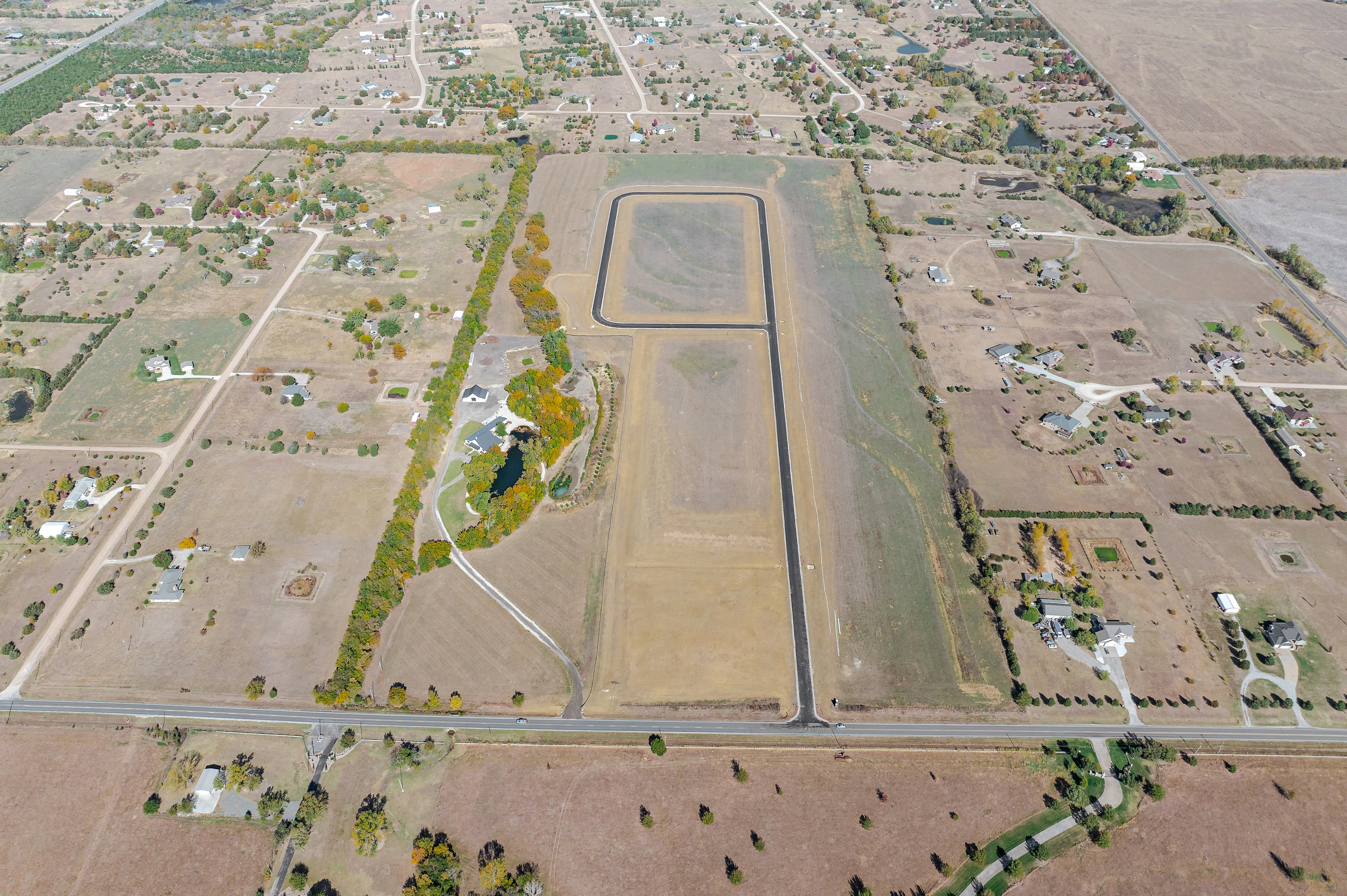 Aerial view of rural farmland with scattered houses, dirt roads, and a paved oval track surrounded by autumn trees.