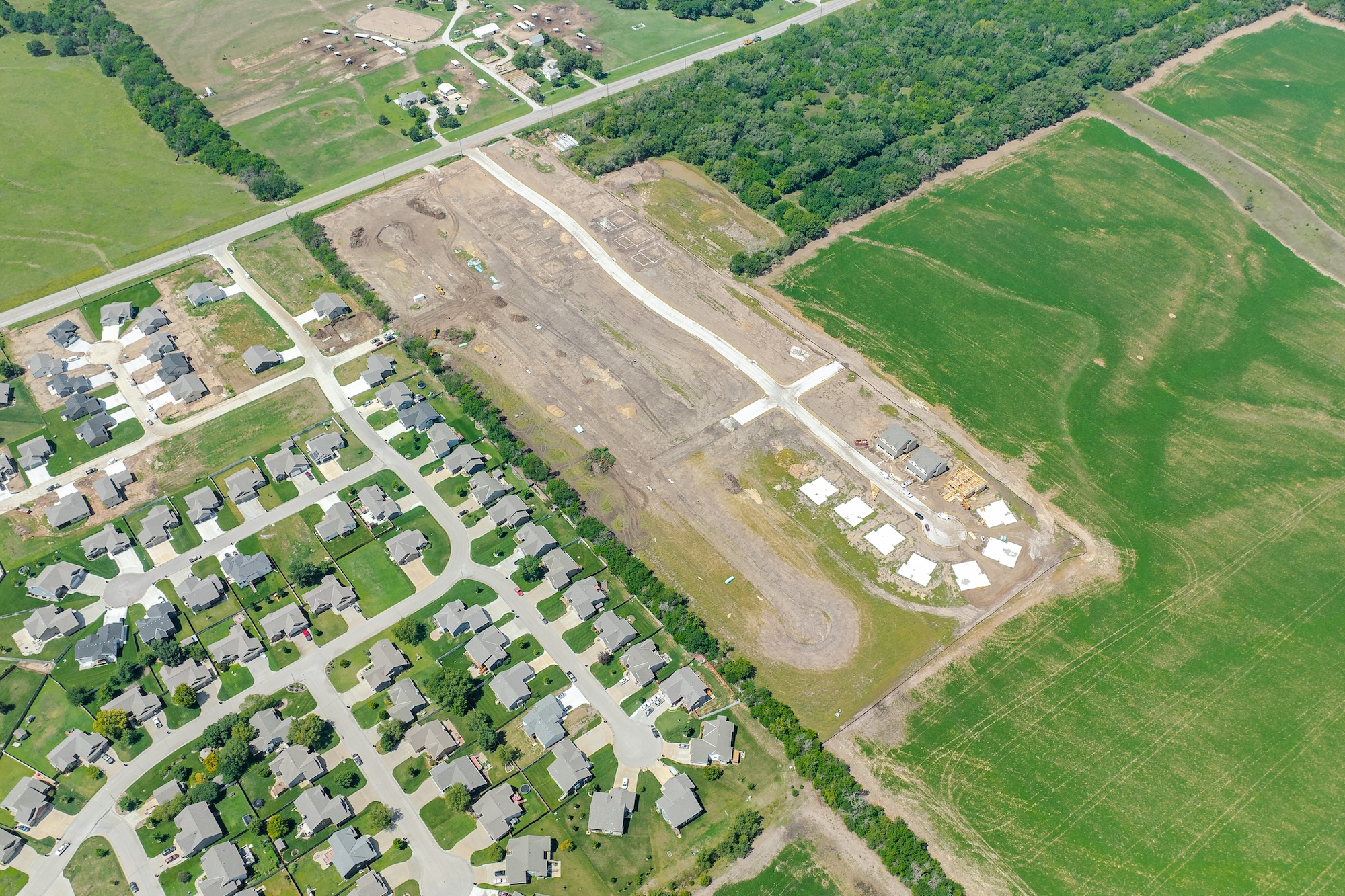 Aerial view of a suburban neighborhood adjacent to a new housing development construction site surrounded by green fields and trees.