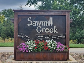 Wooden neighborhood sign reading 'Sawmill Creek' with flower bed of red, white, and pink flowers beneath it.