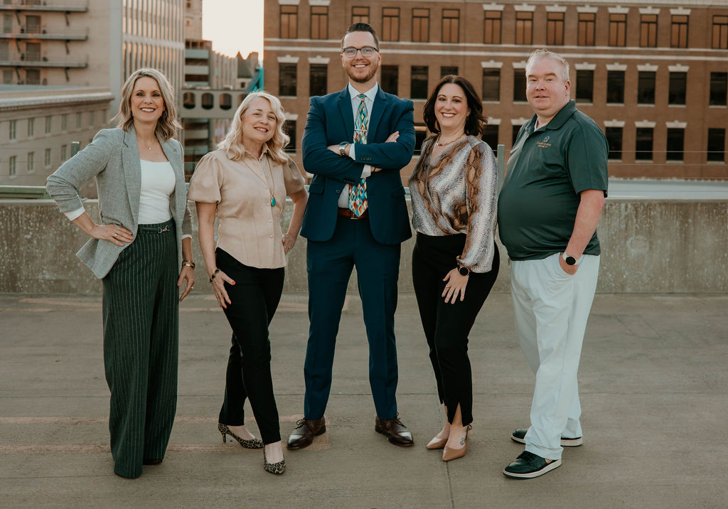 Five professionally dressed people standing outdoors in a city setting, smiling at the camera.