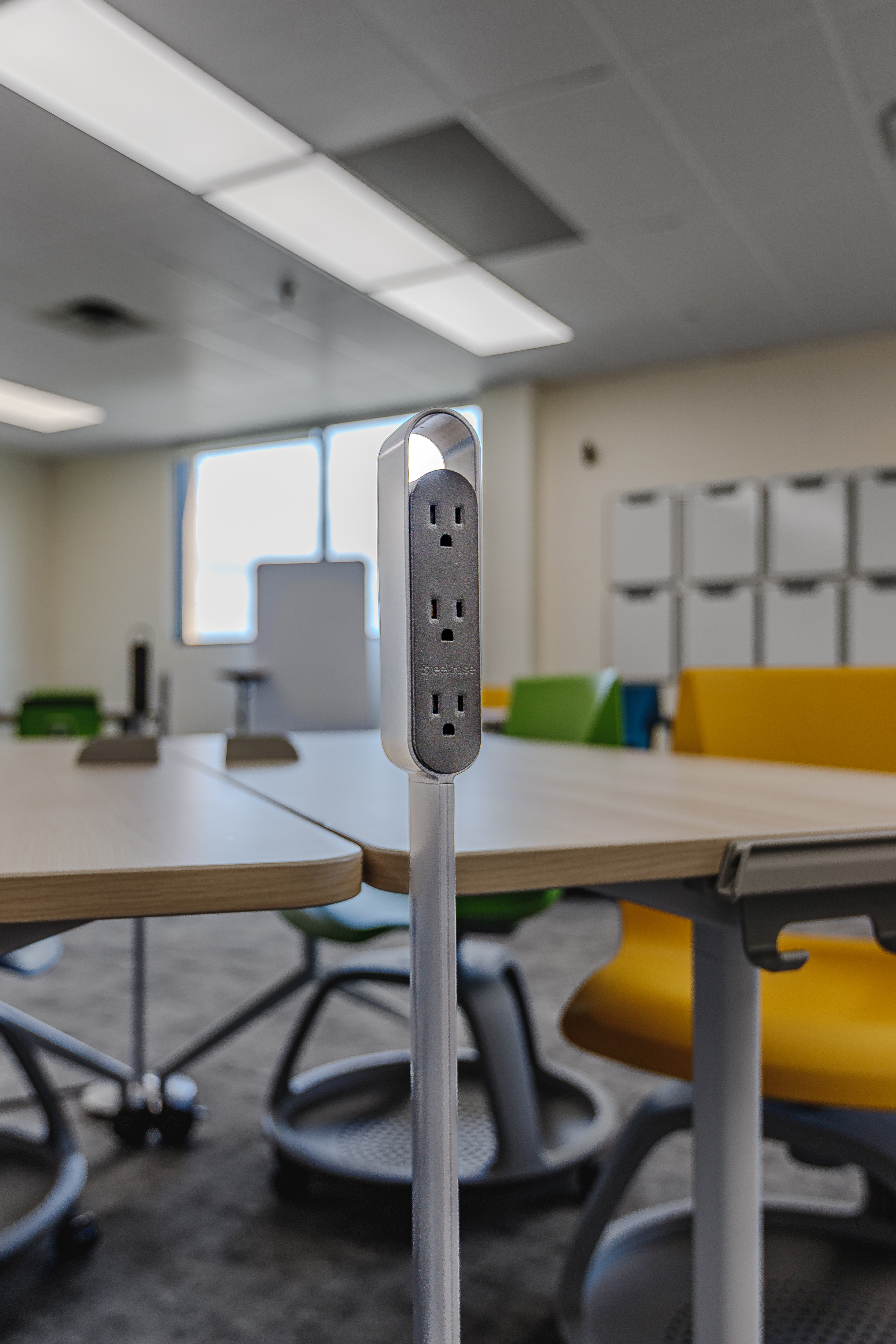 Mobile power strip with three electrical outlets mounted on a pole between desks in a classroom with colorful chairs.