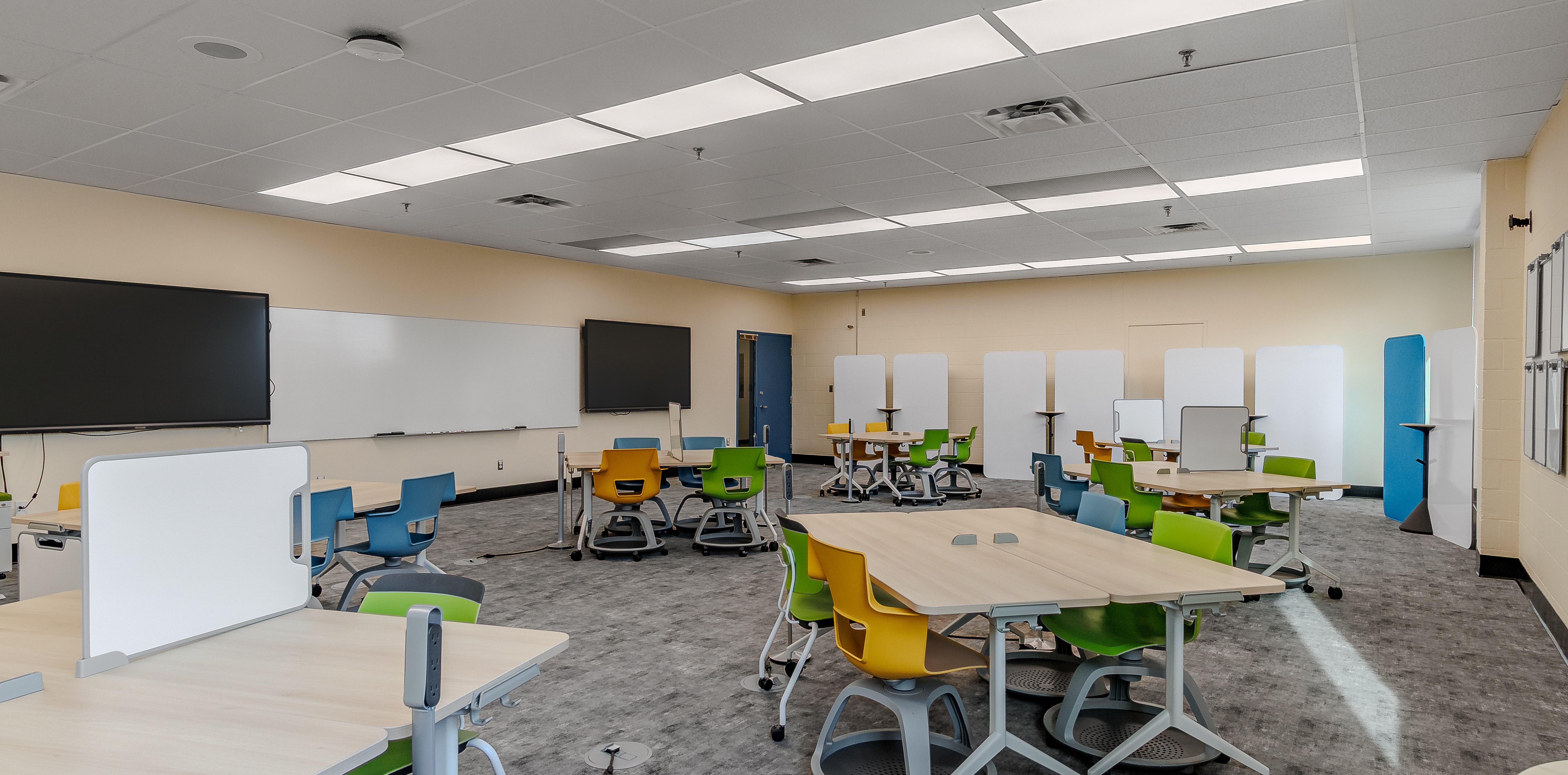Bright classroom with modern tables and multicolored chairs including green, yellow, and blue, featuring whiteboard dividers and two large black monitors on the wall.