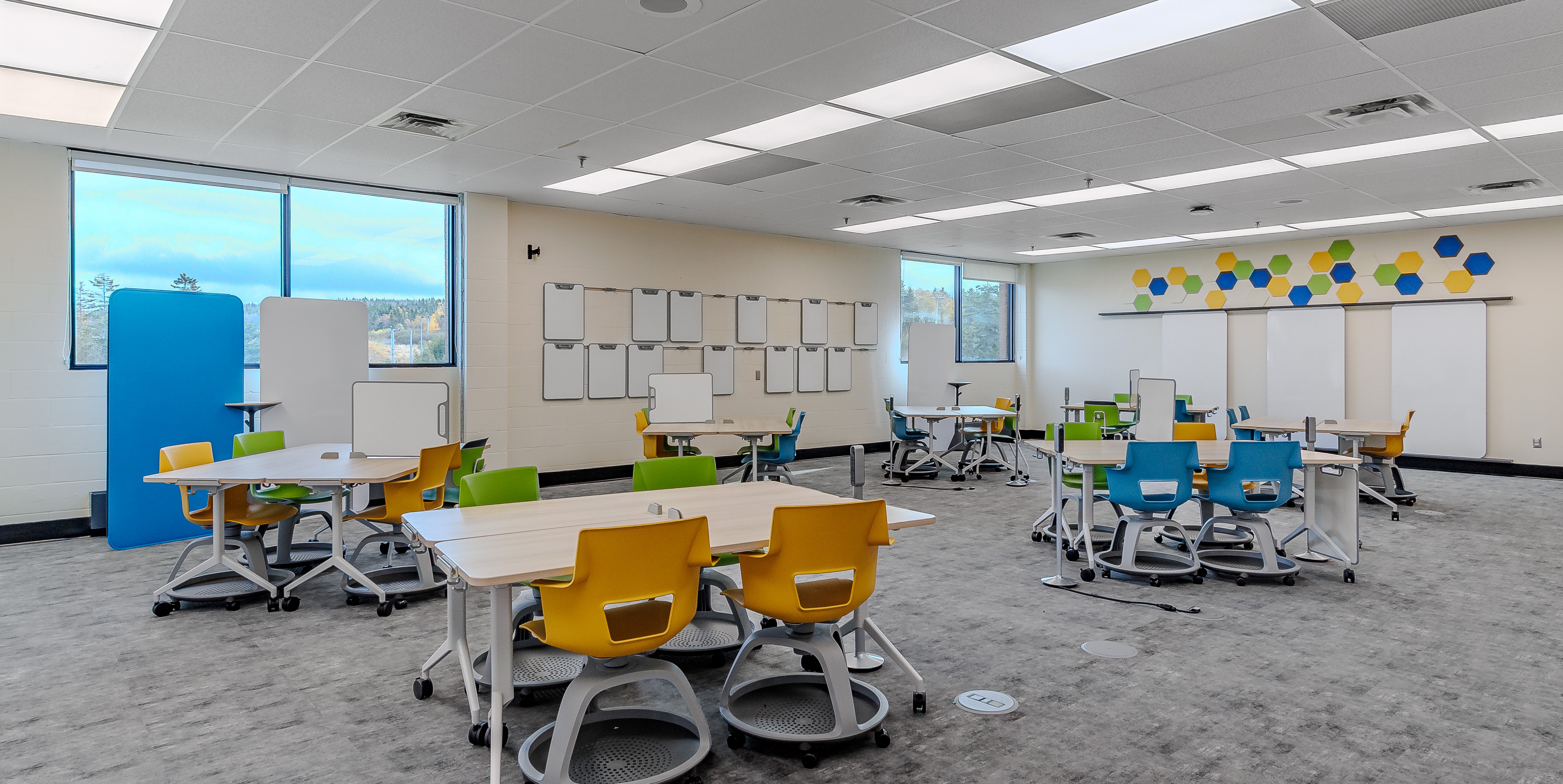 Bright classroom with colorful mobile chairs in yellow, green, and blue around tables with whiteboard dividers, and windows showing an outdoor view.