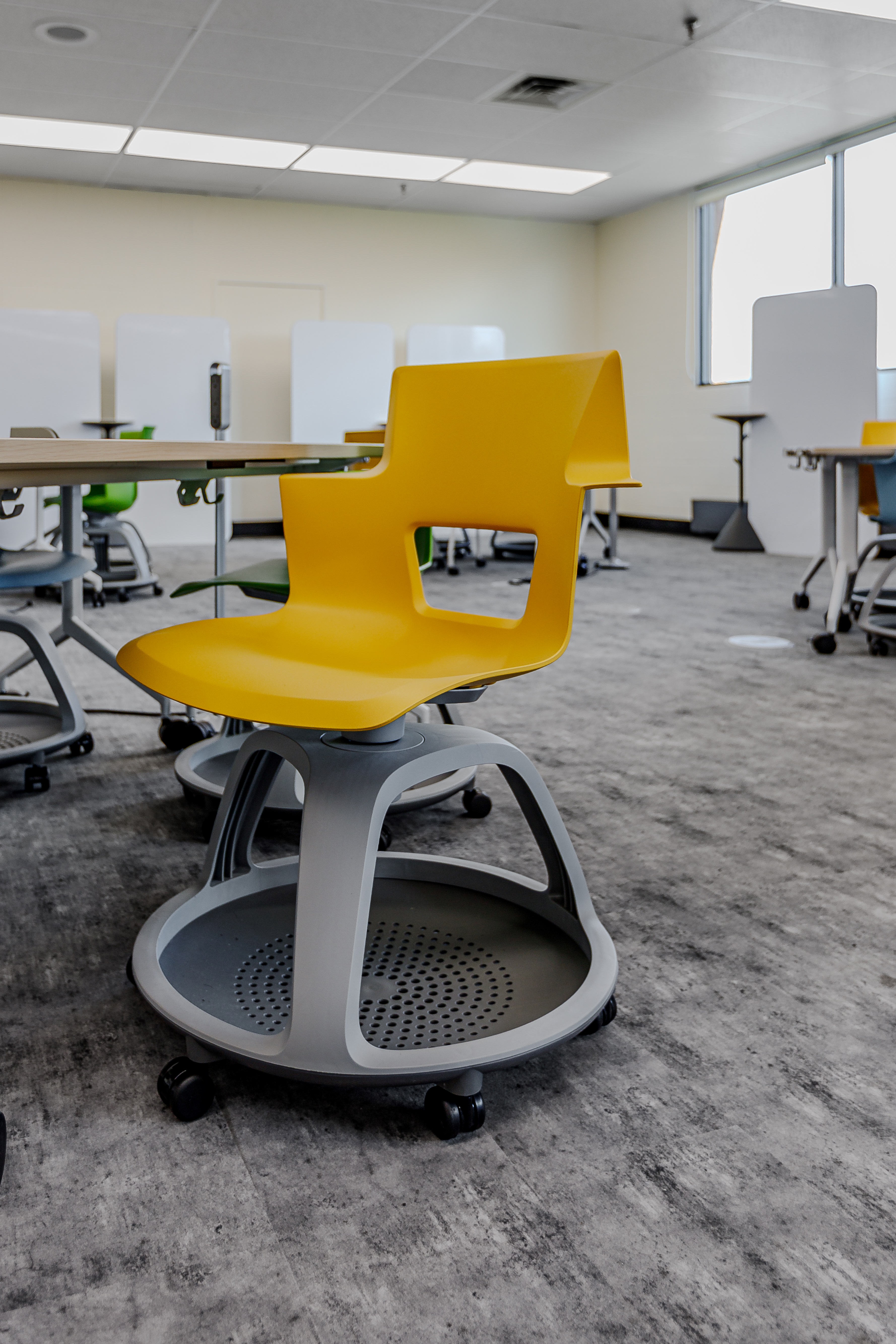 Yellow plastic mobile school chair with storage tray under the seat in a classroom with other colorful chairs and desks.