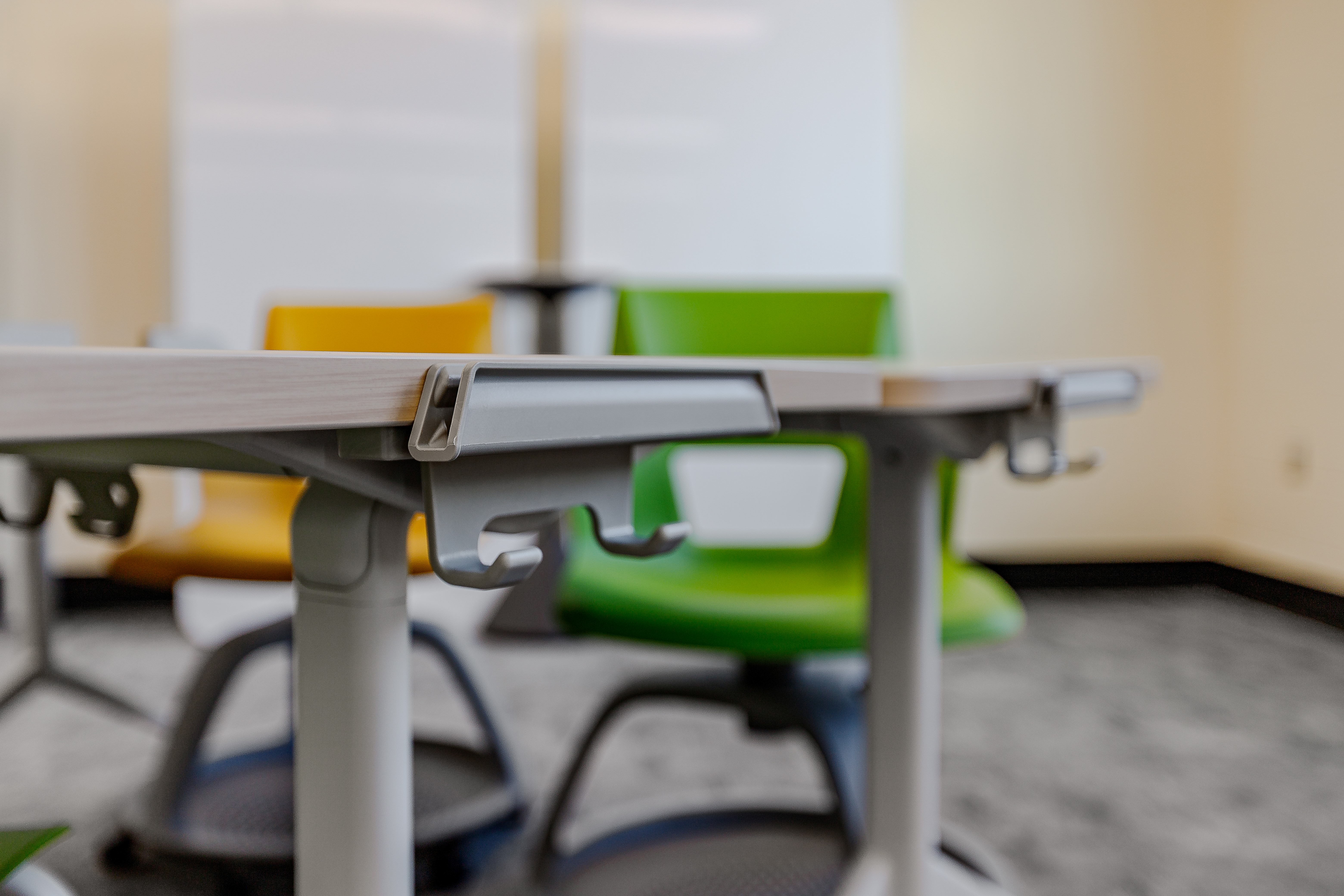 Close-up of a modern classroom table with hooks underneath and colorful chairs in the background.