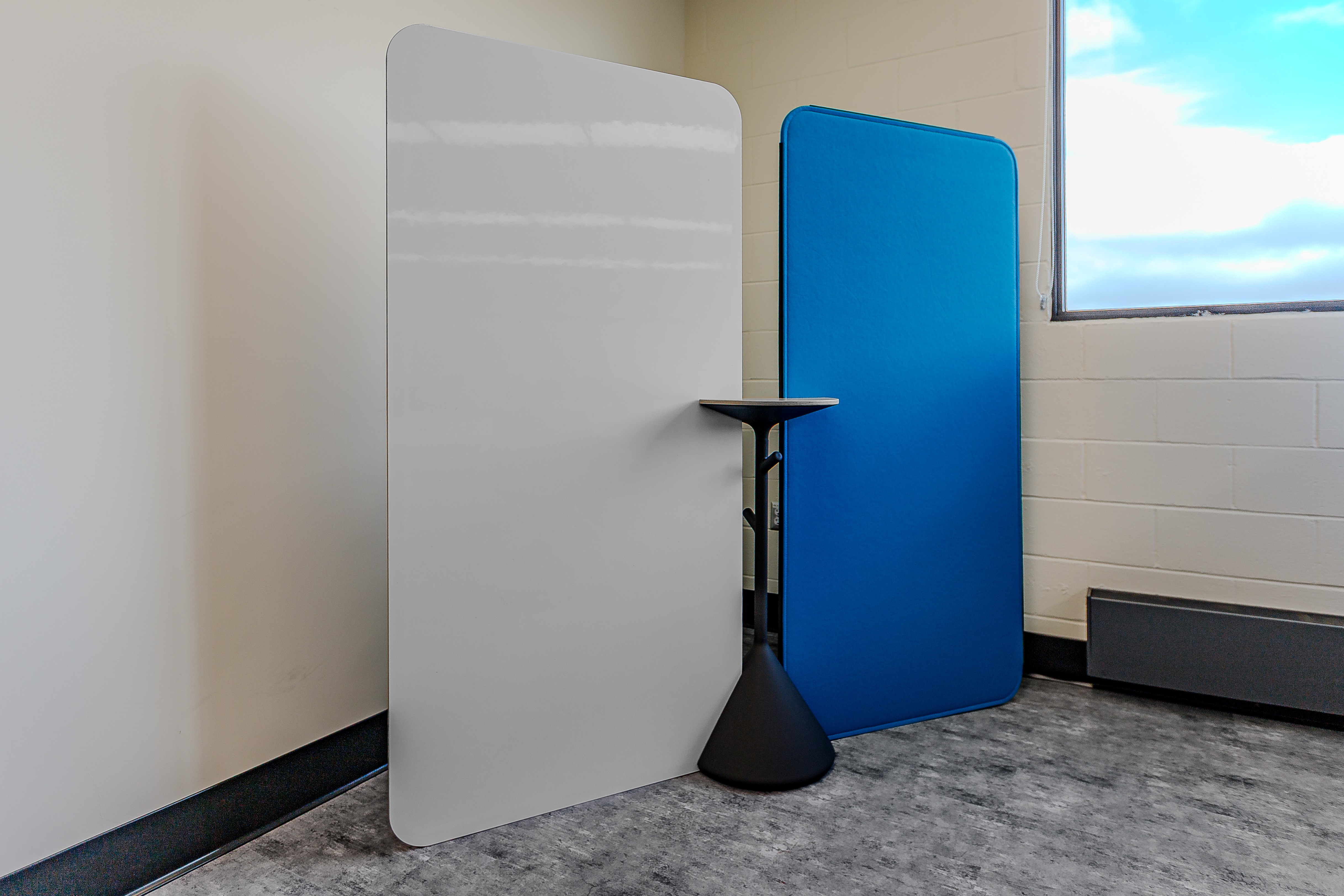 Two modern privacy panels, one white and one blue, with a small black conical table between them in a room with gray flooring and a window.