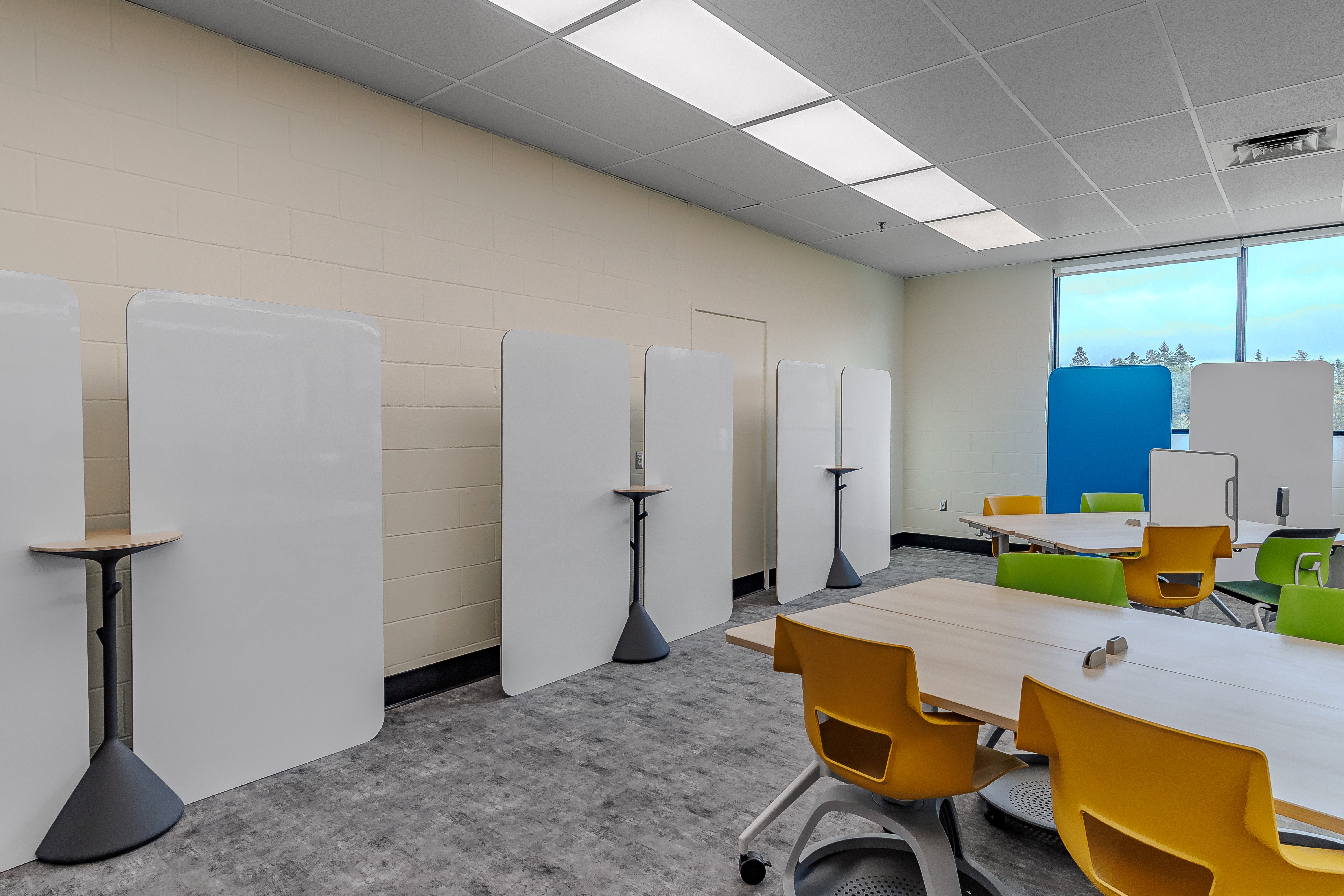Modern classroom with yellow and green chairs, wooden tables, tall white privacy screens, and large windows with outdoor view.