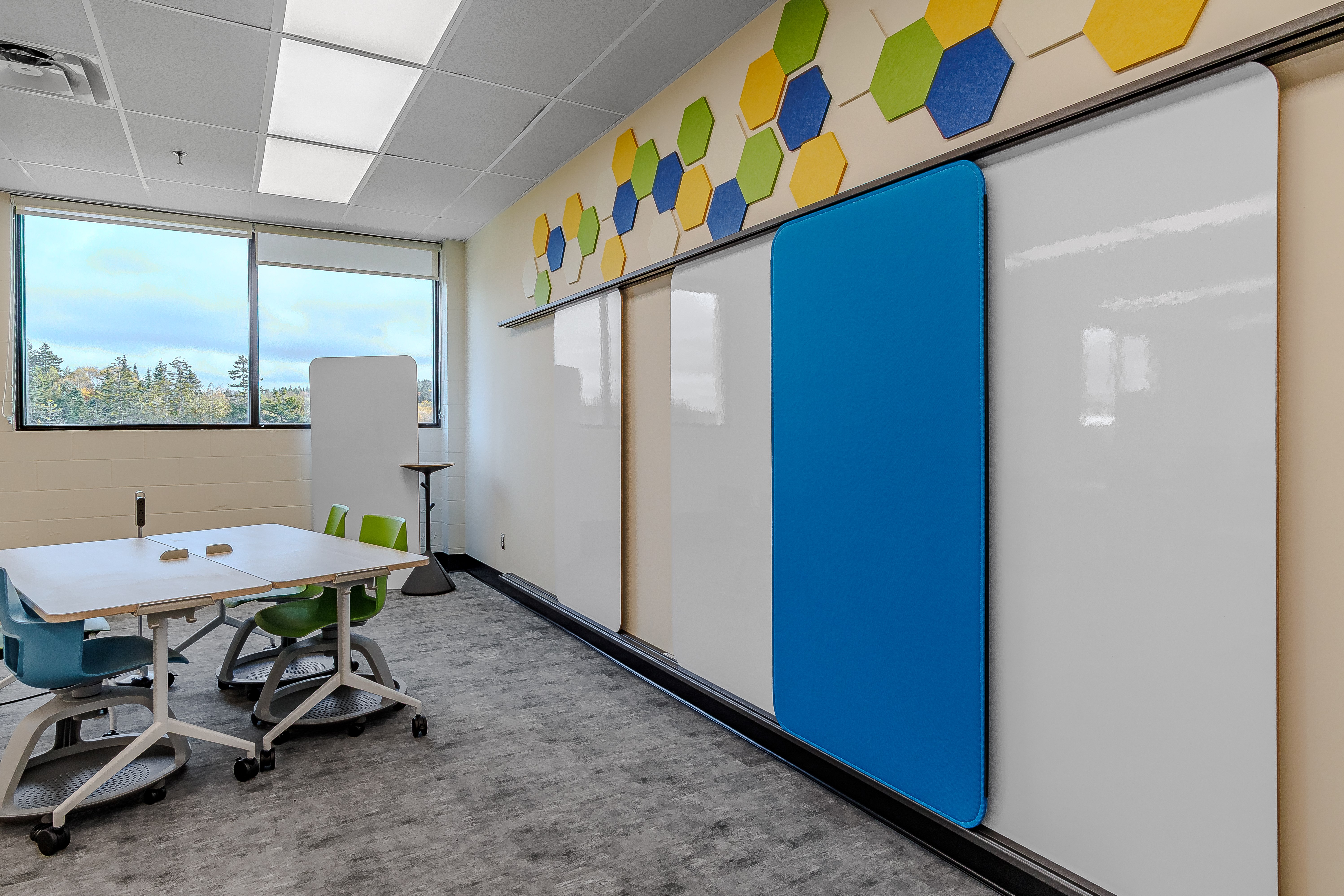 Modern classroom with whiteboards on the wall, blue and green chairs around tables, and large window showing outdoor trees.