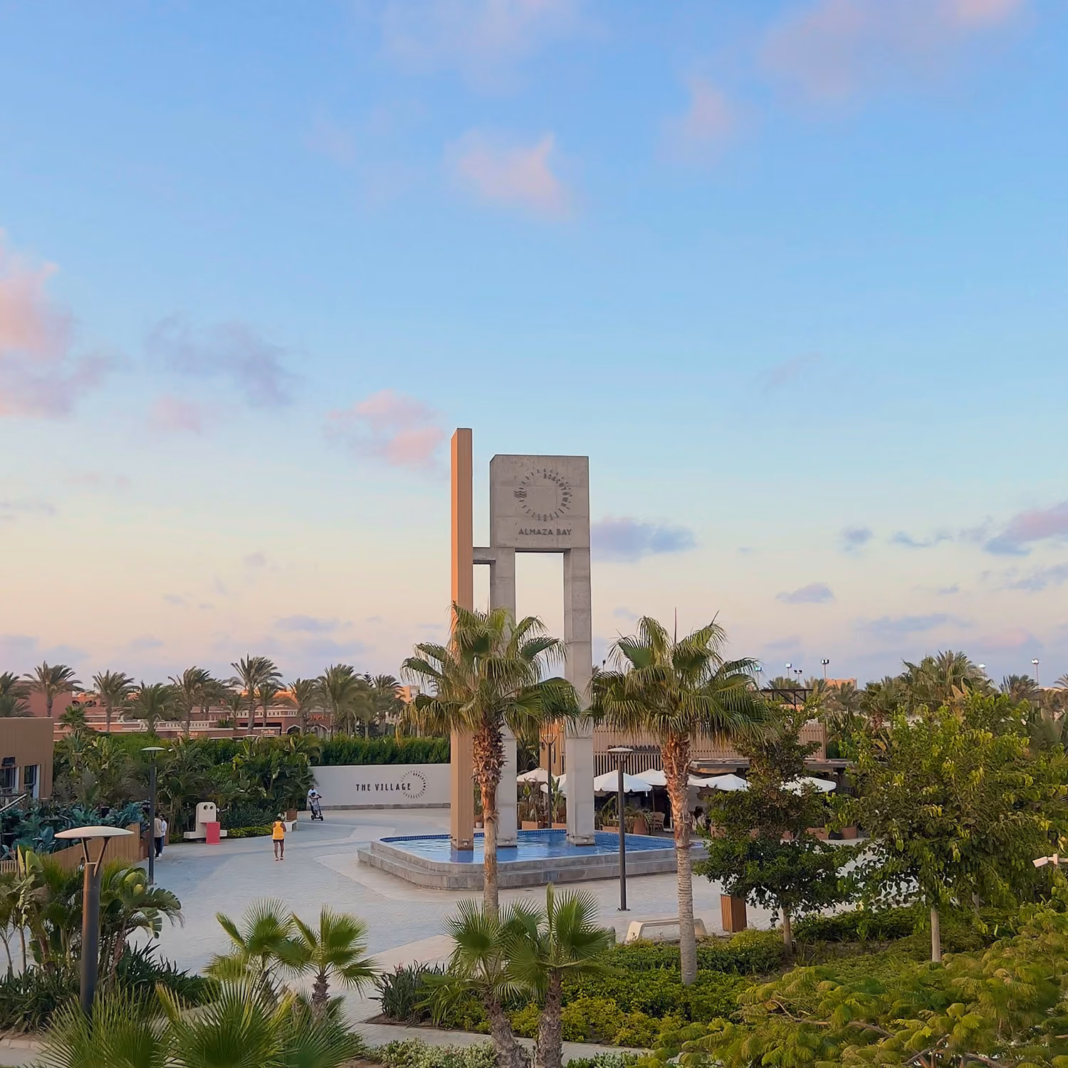 Modern monument with Almaza Bay sign surrounded by palm trees and greenery under a blue sky.