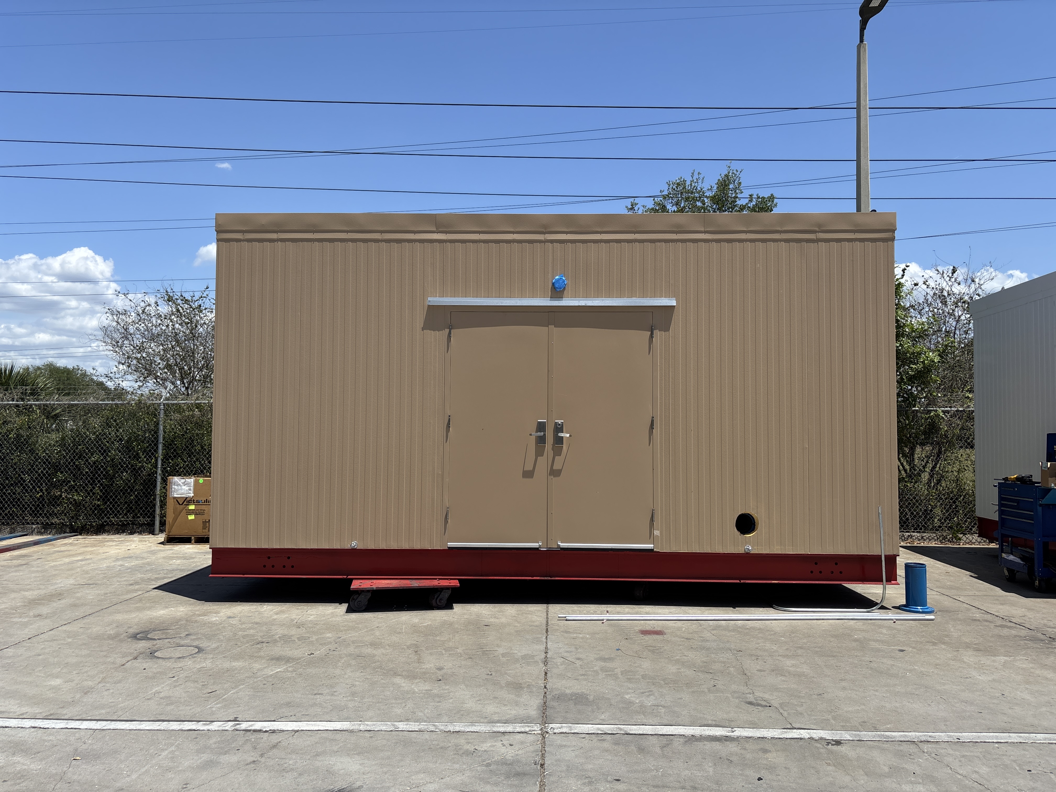 Brown portable building with double doors, red base, on concrete with blue sky background.
