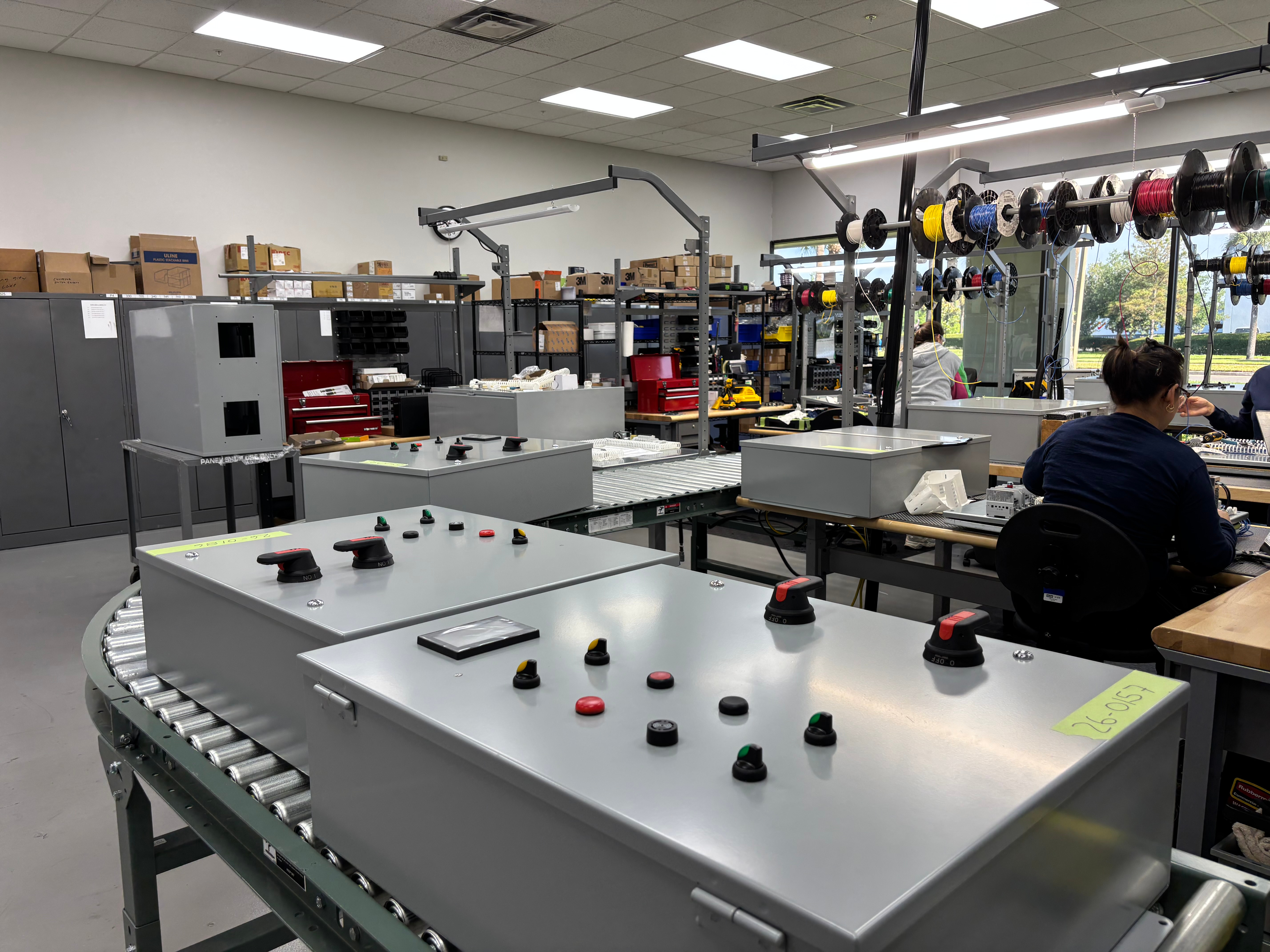 Industrial workspace with control panels on rollers and workers assembling electrical components.