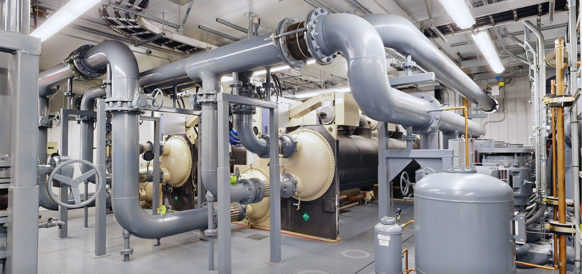 Industrial room with large gray pipes, valves, and machinery in a clean facility.