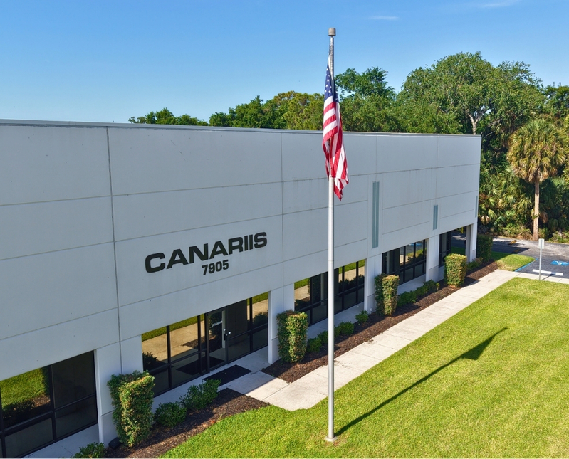 Modern office building with Canariis sign, flagpole with American flag, green lawn, and trees in background.