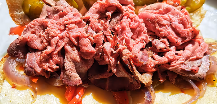 Close-up of thinly sliced rare roast beef on a sandwich with pickles, onions, and red peppers.