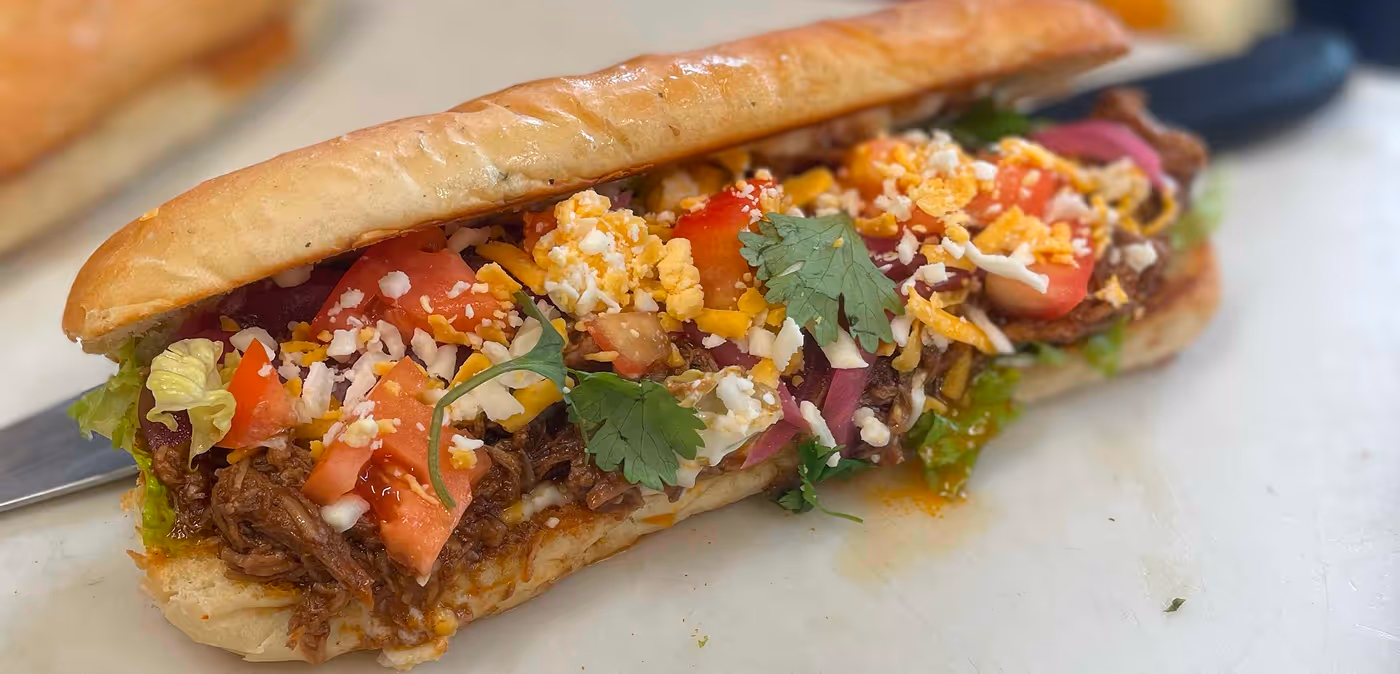 Sandwich with shredded beef, chopped tomatoes, lettuce, cilantro, and shredded cheese on a toasted sub roll.