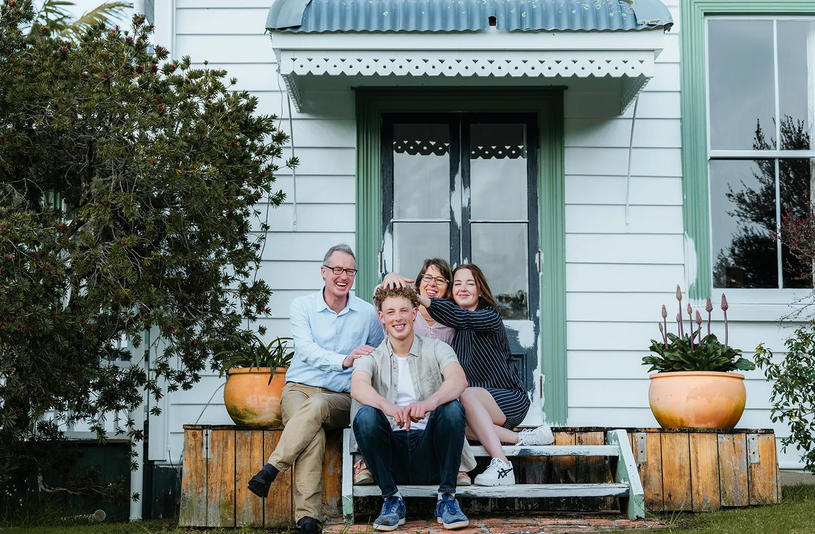 Smiling family of four sitting on wooden steps of a weathered house with plants in terracotta pots on either side.
