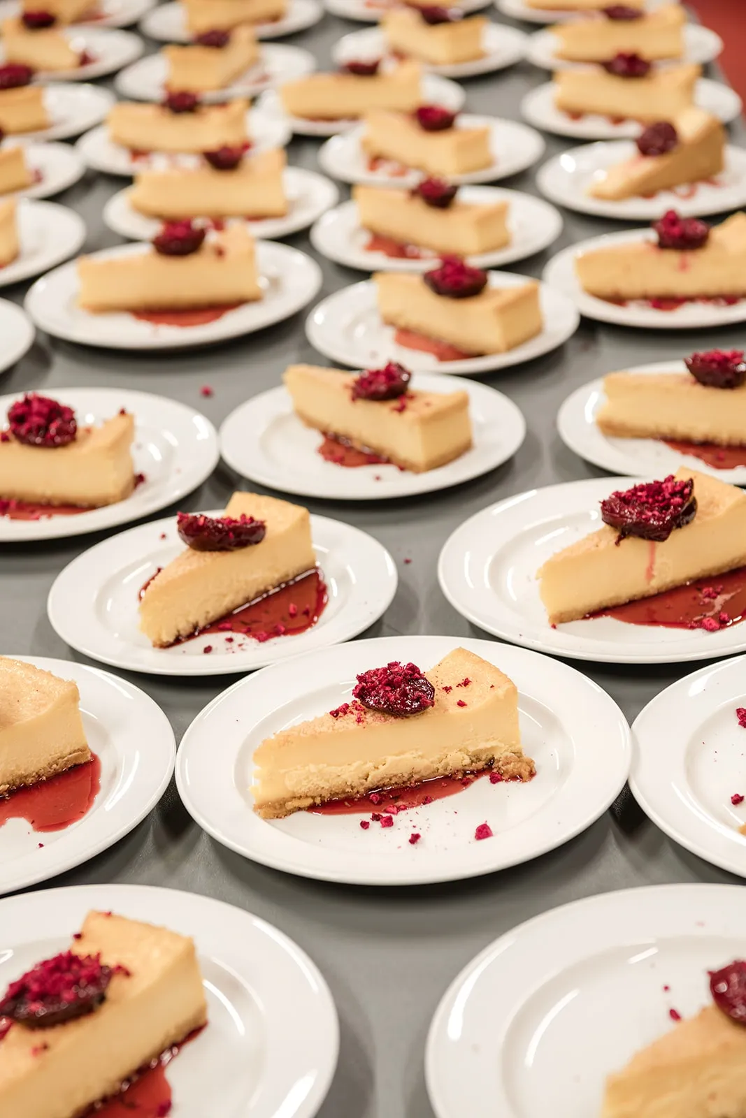 Multiple white plates each with a slice of cheesecake topped with a red berry and red sauce on a grey table.