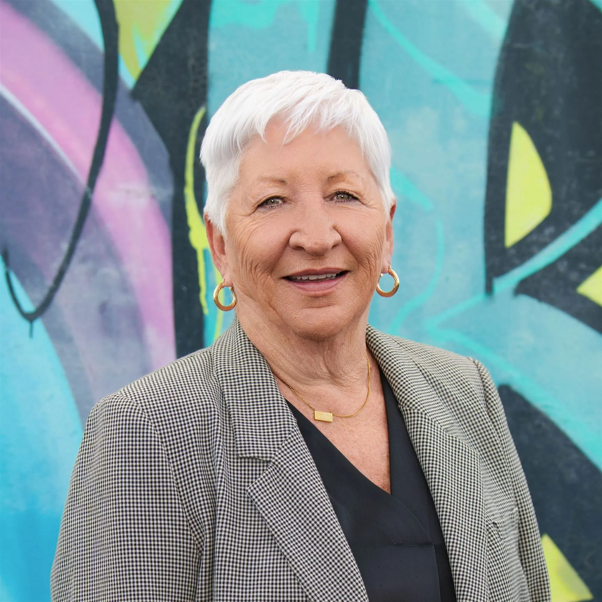 Smiling older woman with short white hair wearing a black and white checkered blazer, gold hoop earrings, and a gold necklace, standing in front of a colourful graffiti wall.