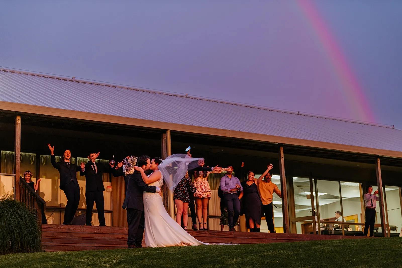 Bride and groom kissing outdoors in front of a modern building with wedding guests cheering on a porch and a rainbow in the sky.