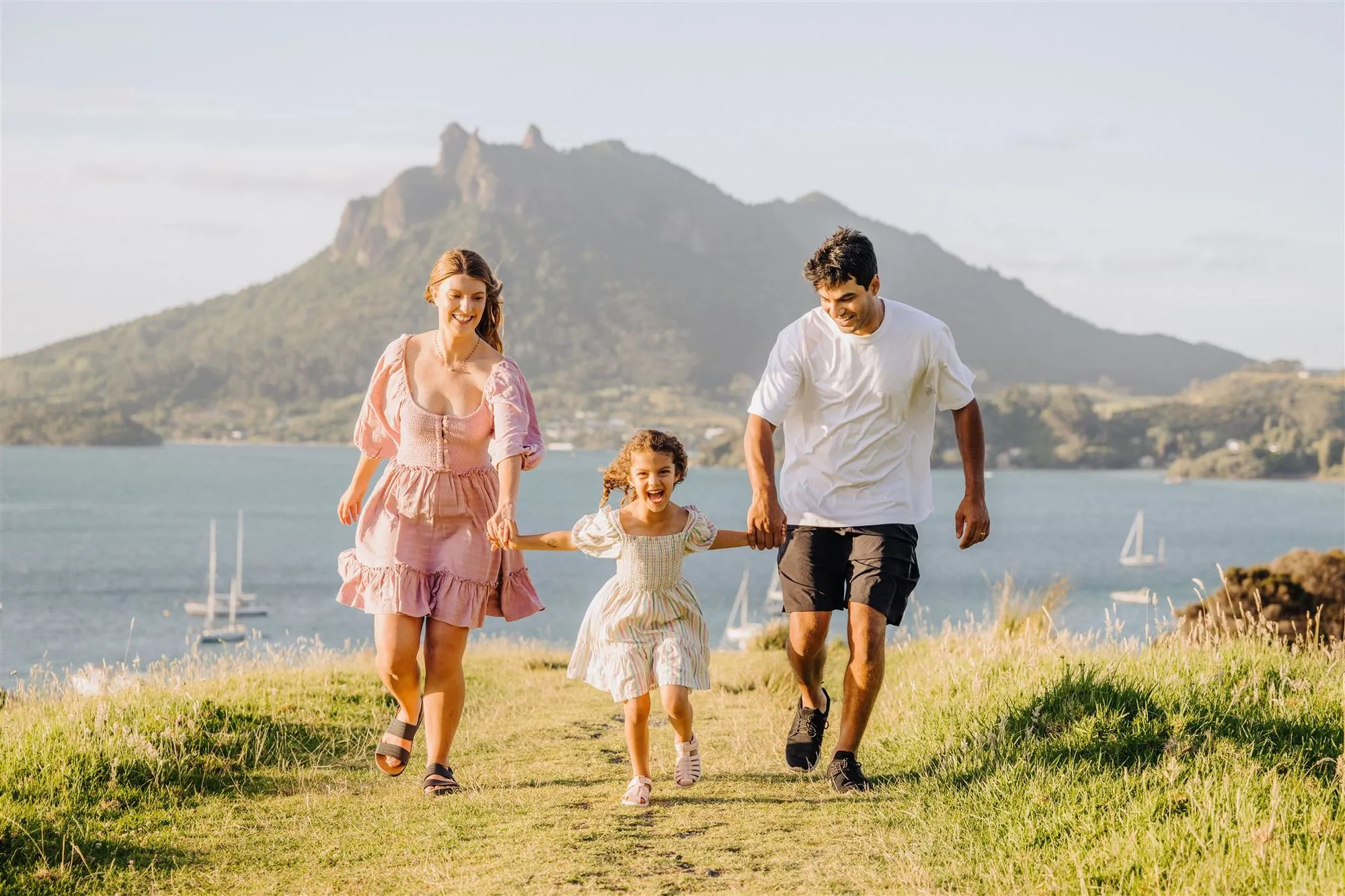Smiling family of three holding hands, walking along a grassy path with a mountain and boats on the water in the background.
